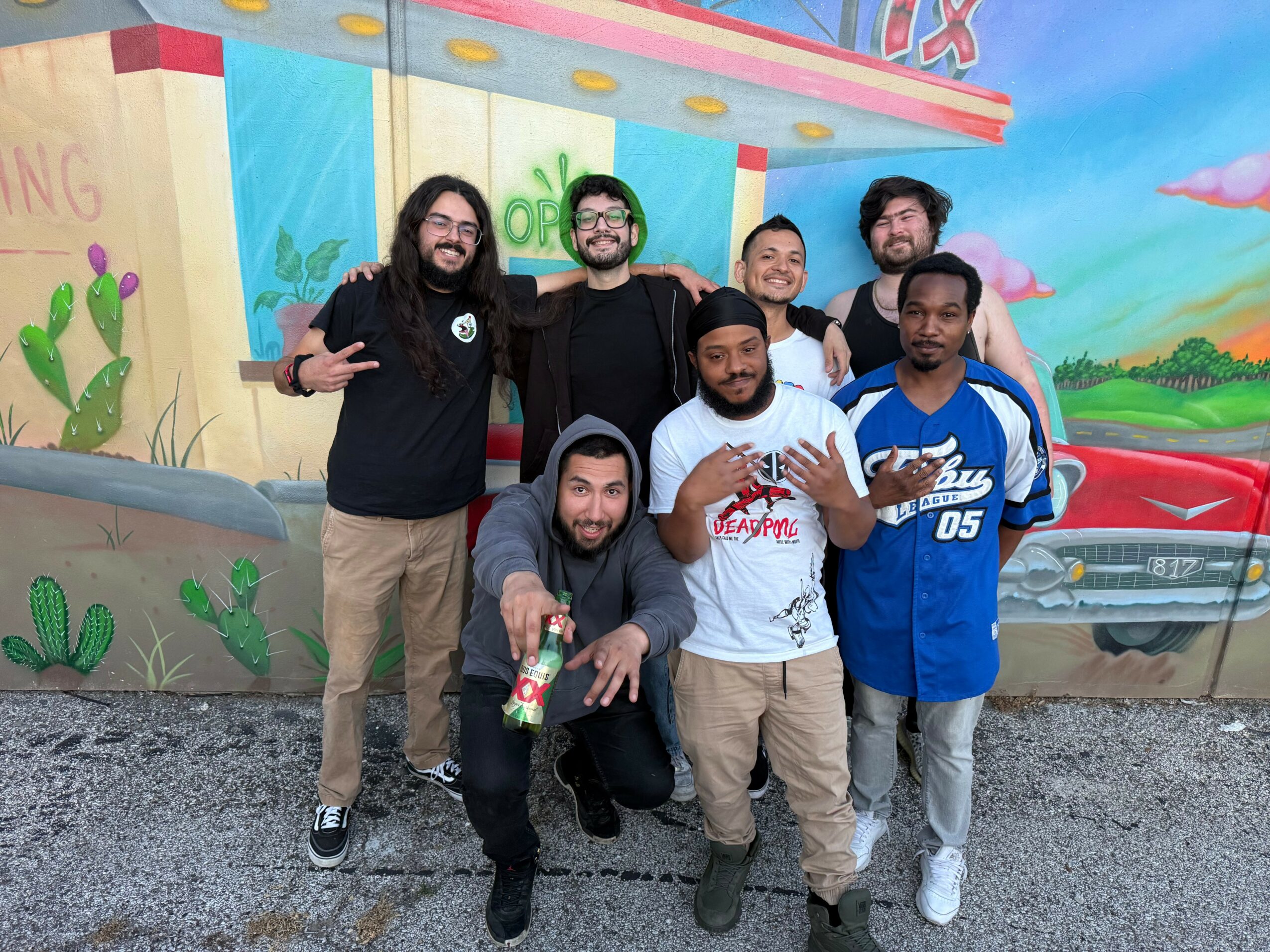 Group of seven men posing in front of a colorful mural with a cartoonish scene, some making hand gestures, smiling.