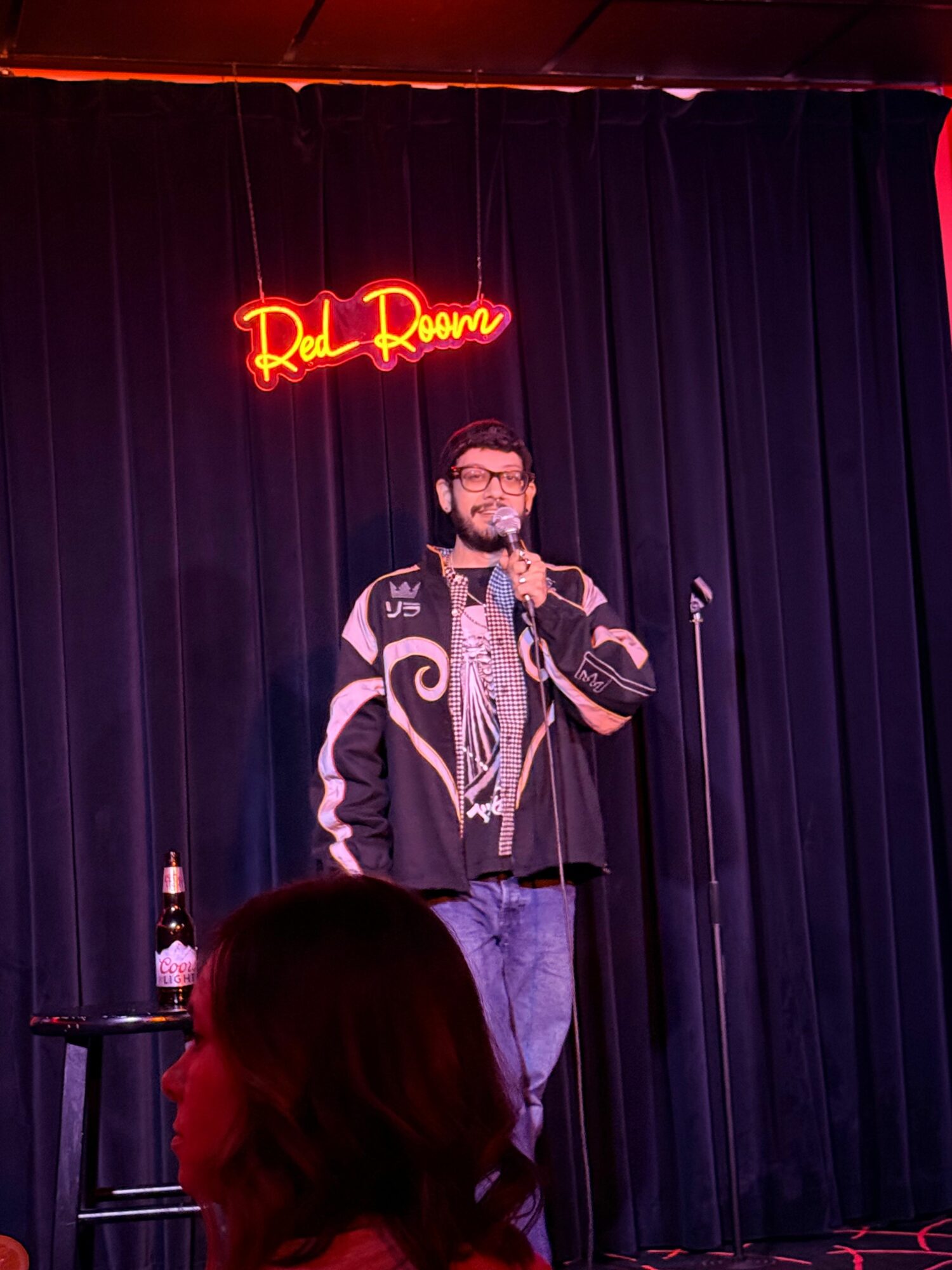 Person standing on stage with a microphone, wearing a jacket with designs, in front of a black curtain and red neon sign