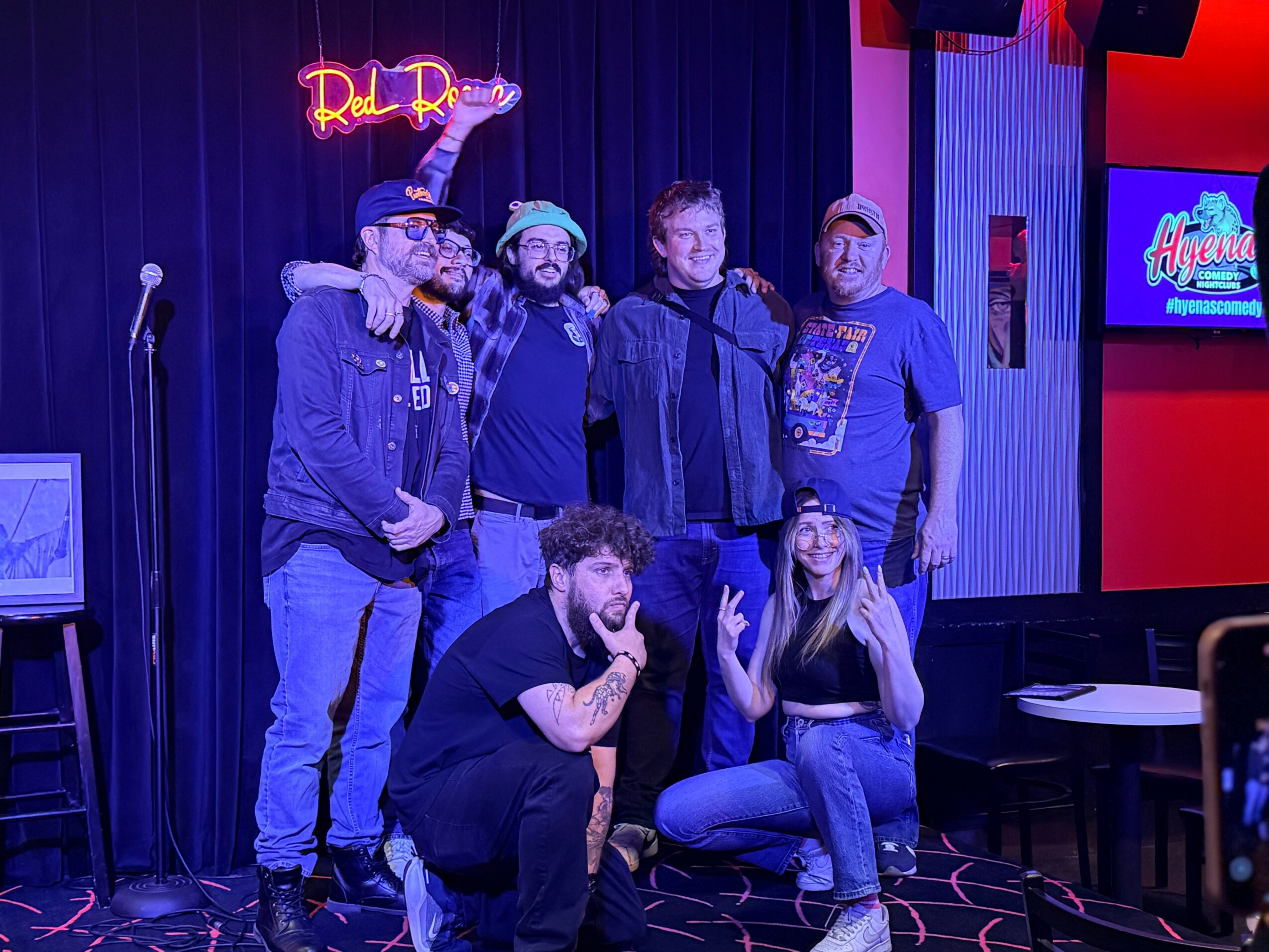 Group of six people on stage, some standing and some kneeling, with a neon sign and a screen in the background.