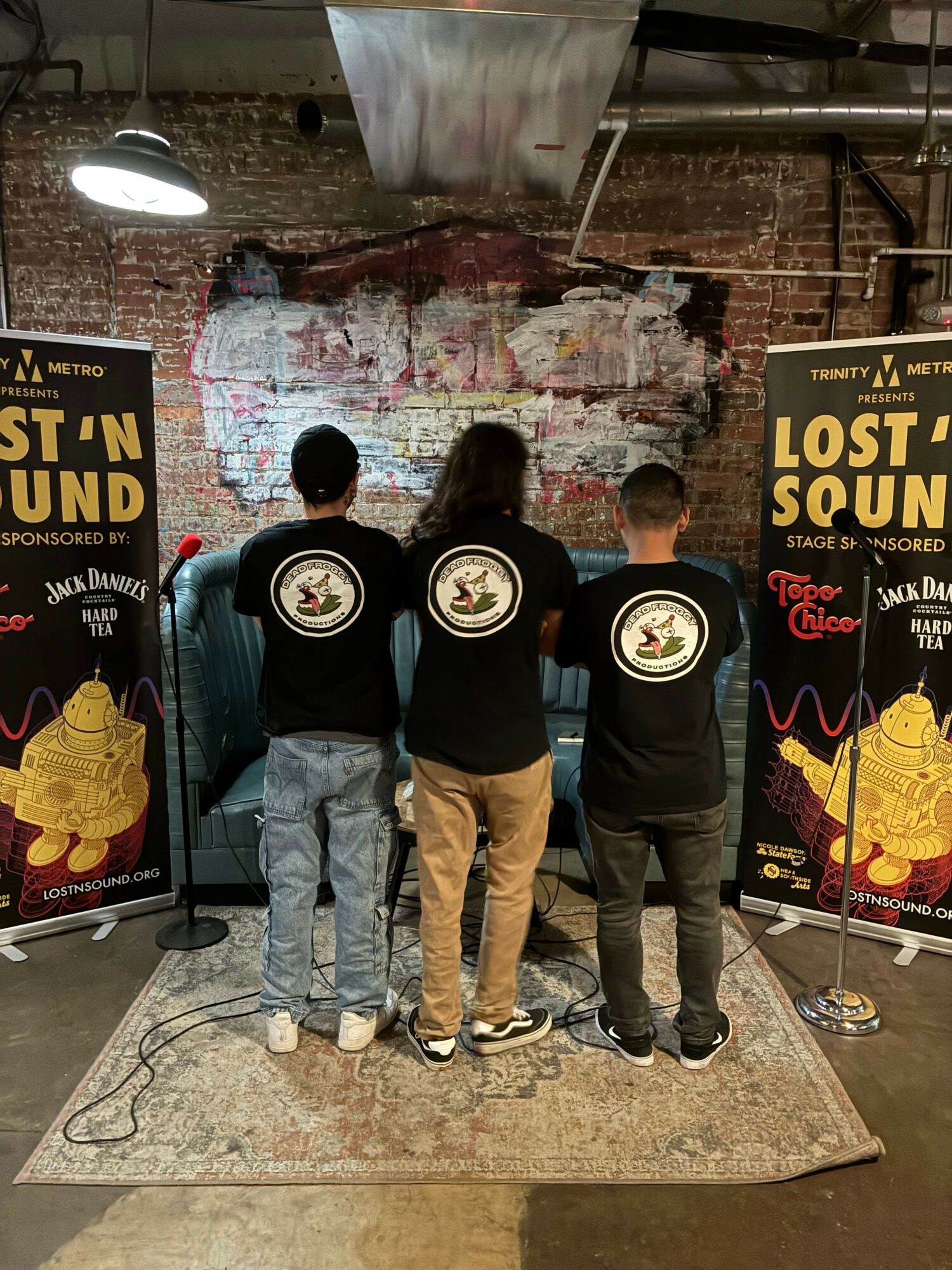 Three people stand on a stage with a microphone, in front of a backdrop and posters, in an indoor setting.