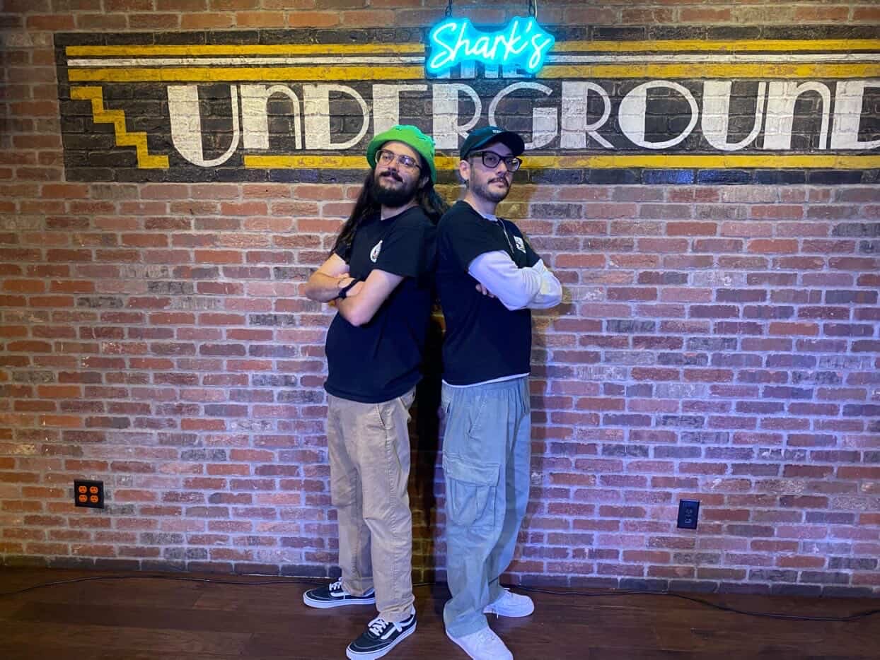 Two men stand back-to-back in front of a brick wall with a neon sign and underground sign, arms crossed, looking at camera.