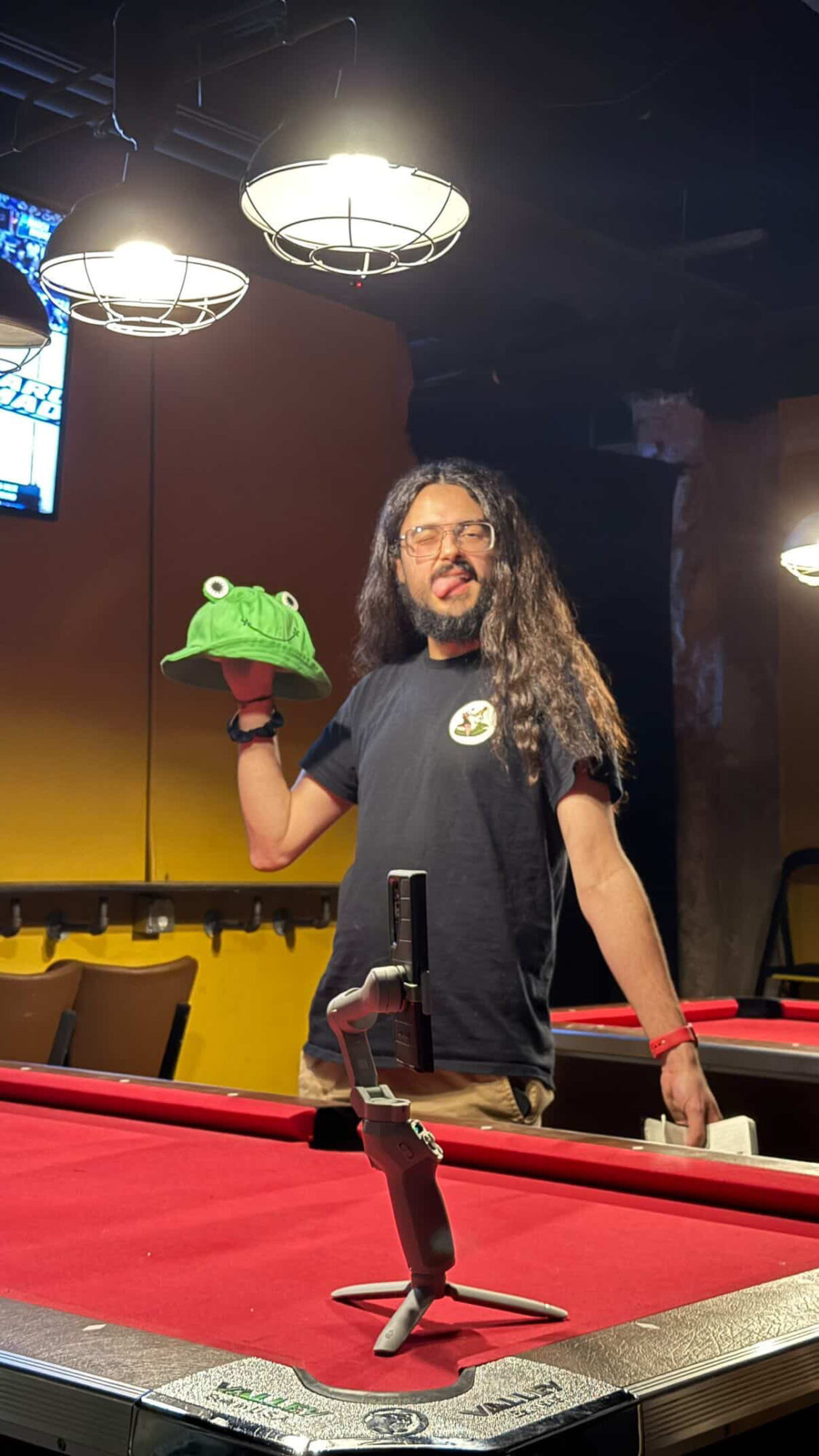 Person with long hair and glasses holding a green frog hat in a game room with pool tables and hanging lights