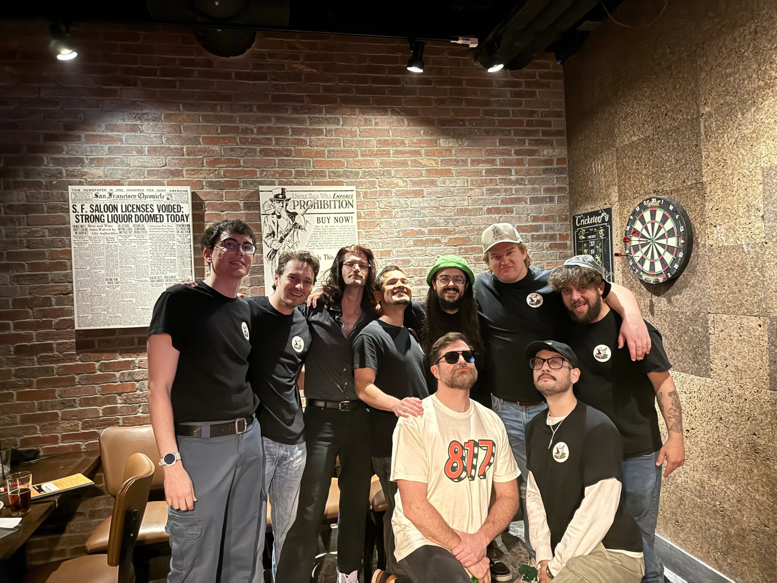 Group of nine people standing together in a room with brick and stone walls, some wearing sunglasses and casual clothing.