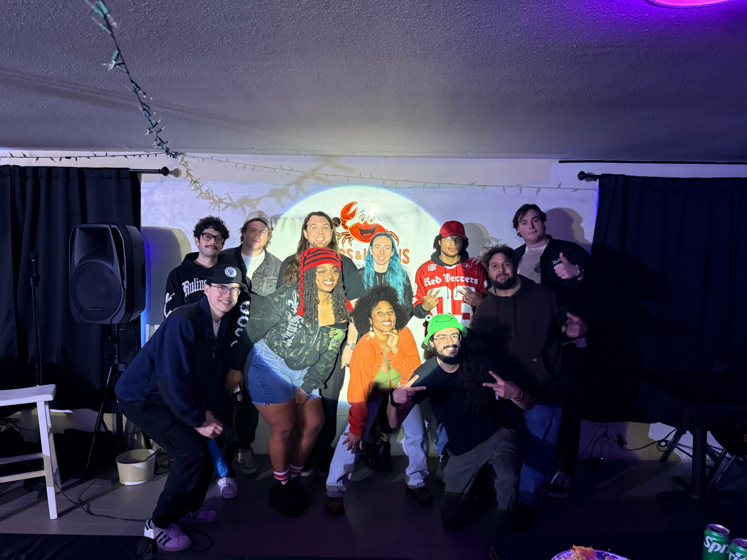 Group of people posing on stage with a festive background, some wearing costumes and accessories, in a dimly lit room.