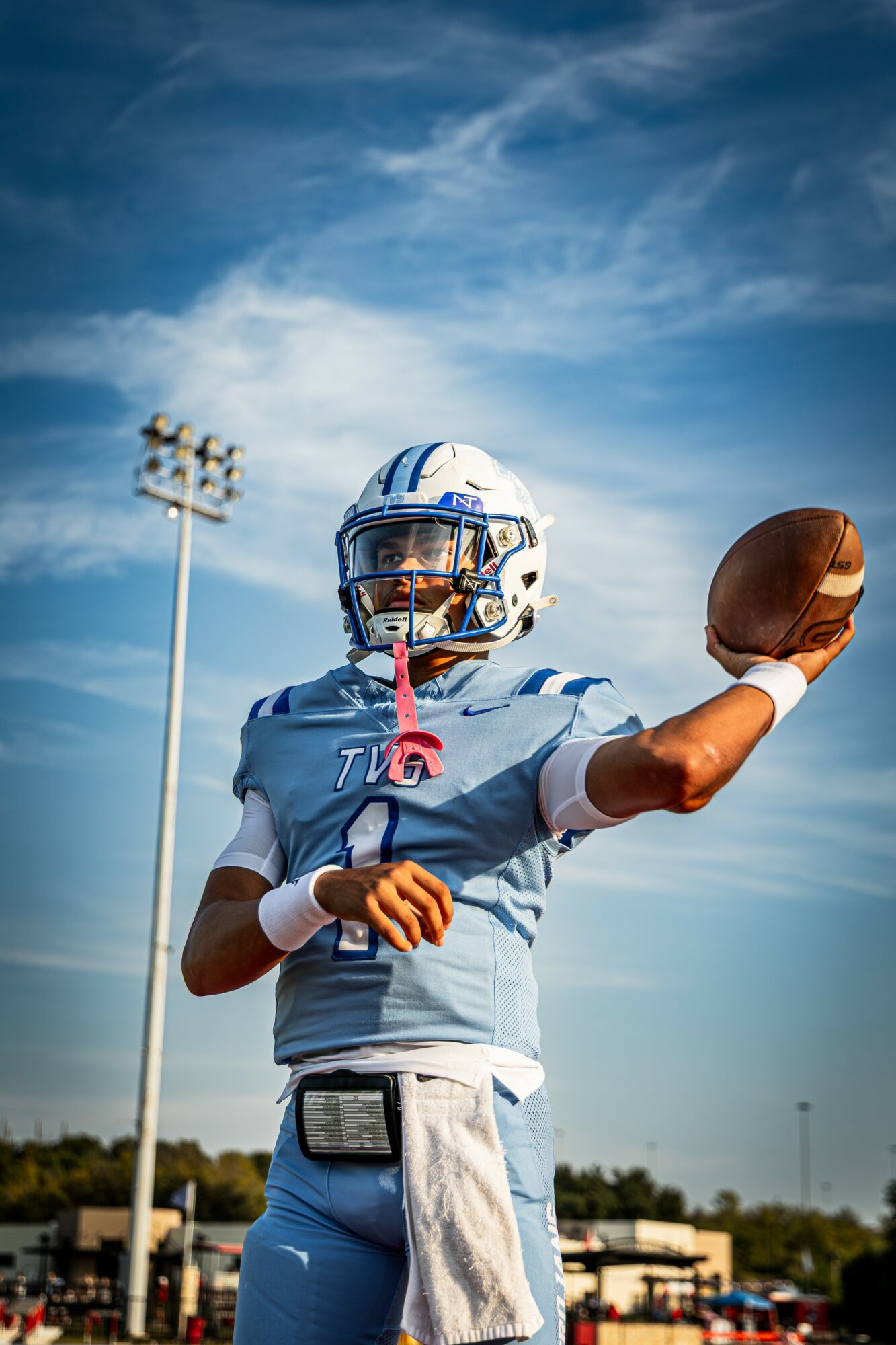 Football player in uniform holding a football against a blue sky with clouds, stadium lights, and trees in background.