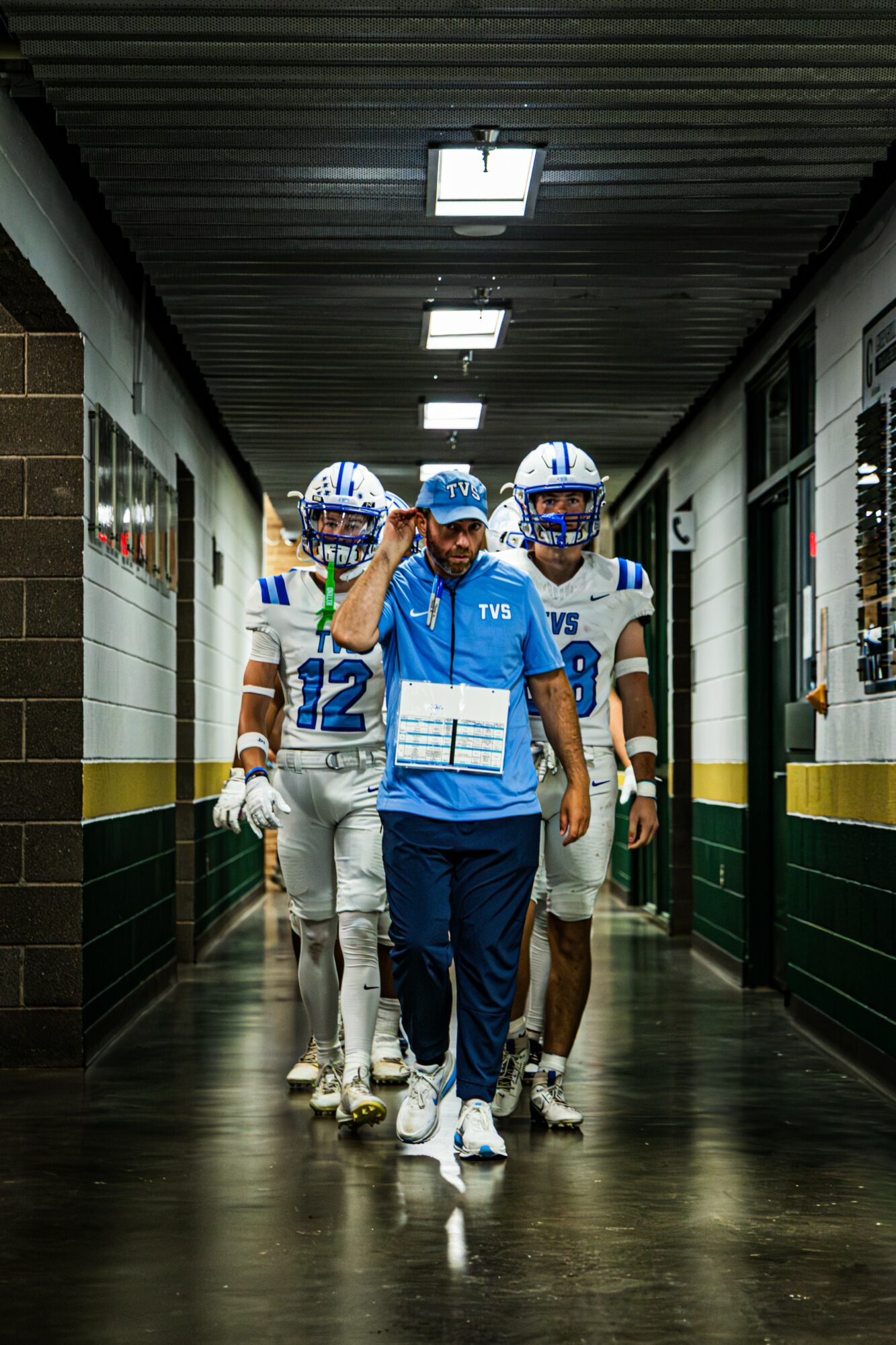 Four football players walking down a hallway, wearing helmets and uniforms, with one player in front waving.
