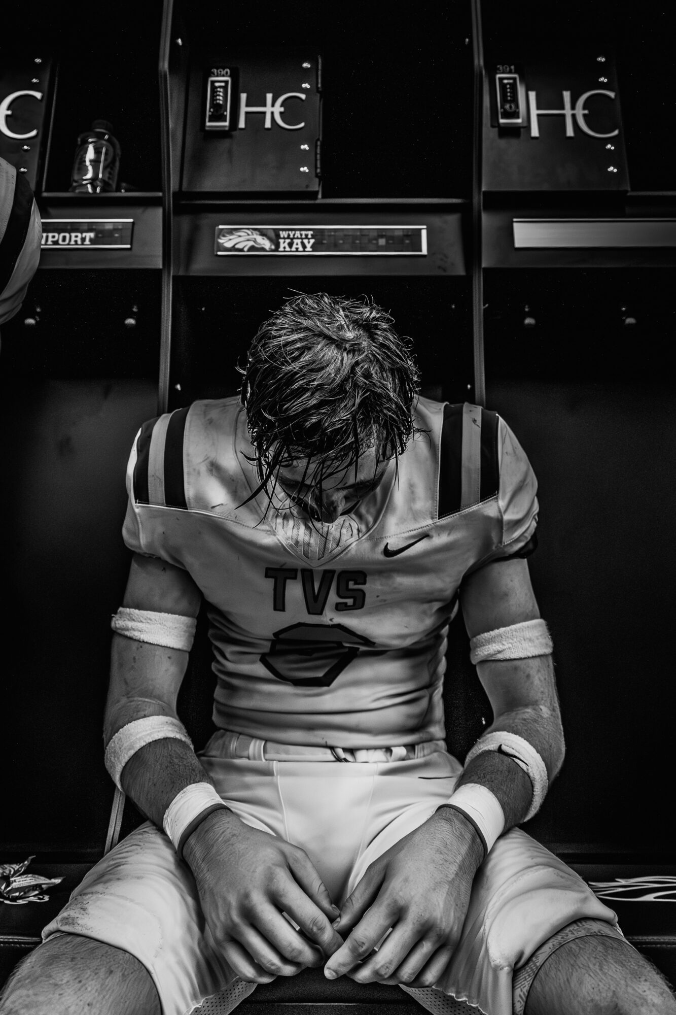 Football player sitting in locker room with head down, wearing uniform and wristbands, lockers in background.