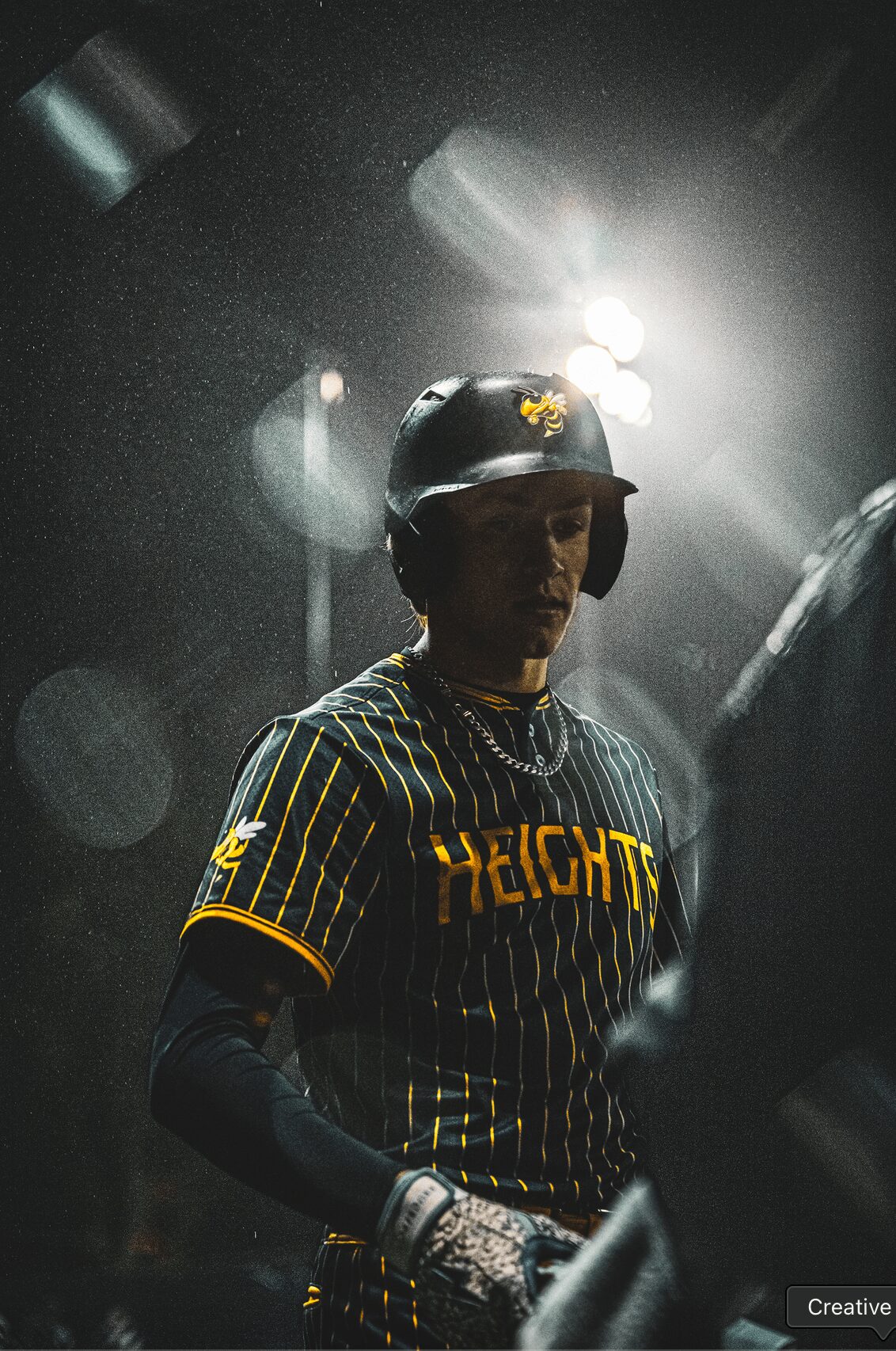 Person in a baseball uniform and helmet, standing in a dark environment with bright light behind, partially obscured face.