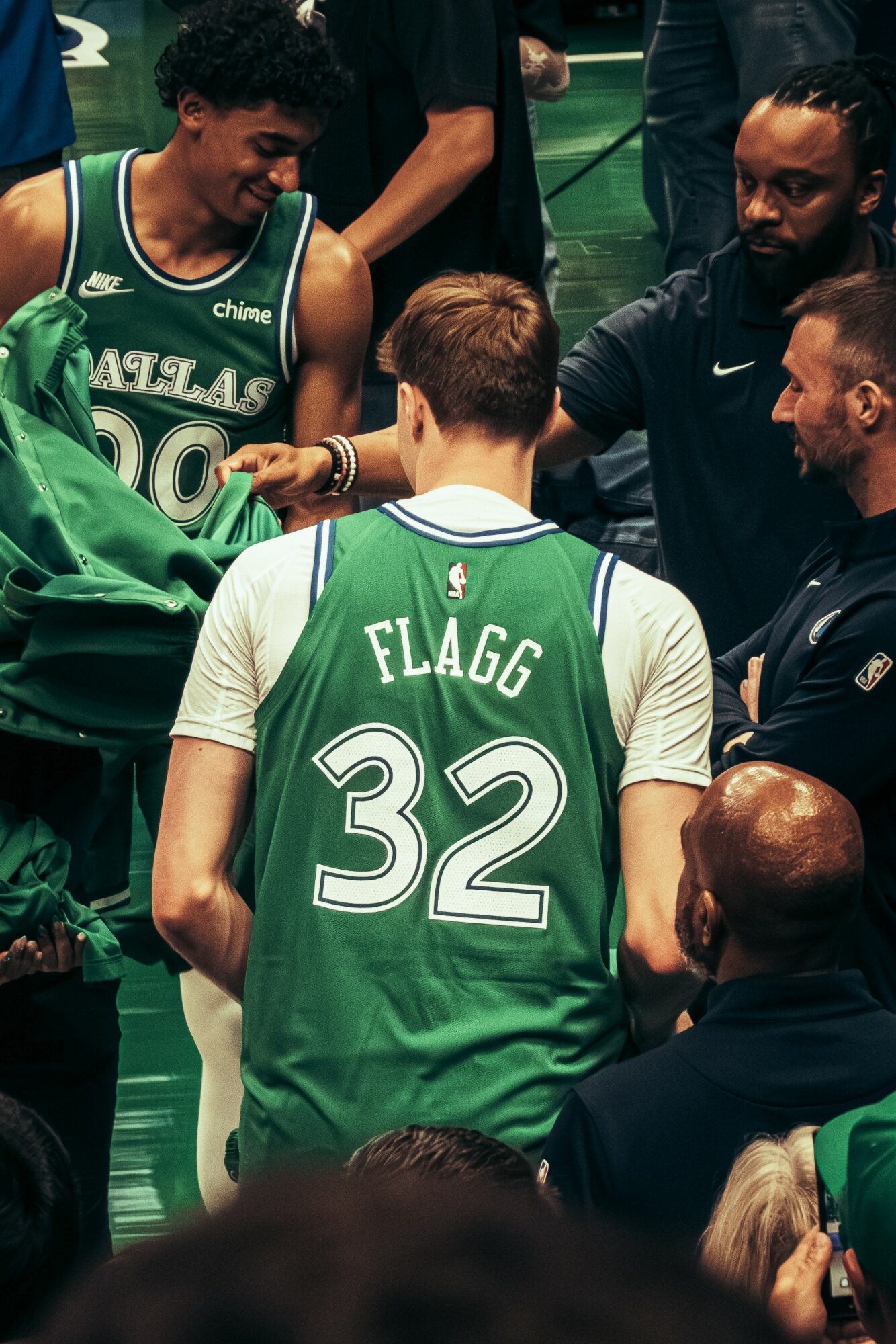 Group of basketball players and coaches in green jerseys and black clothing on a court, engaging in discussion.