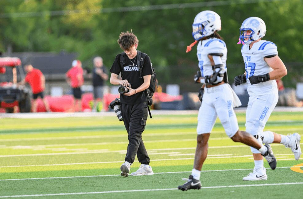 A young person on a football field holding a camera, with two football players running nearby, and spectators in the background.