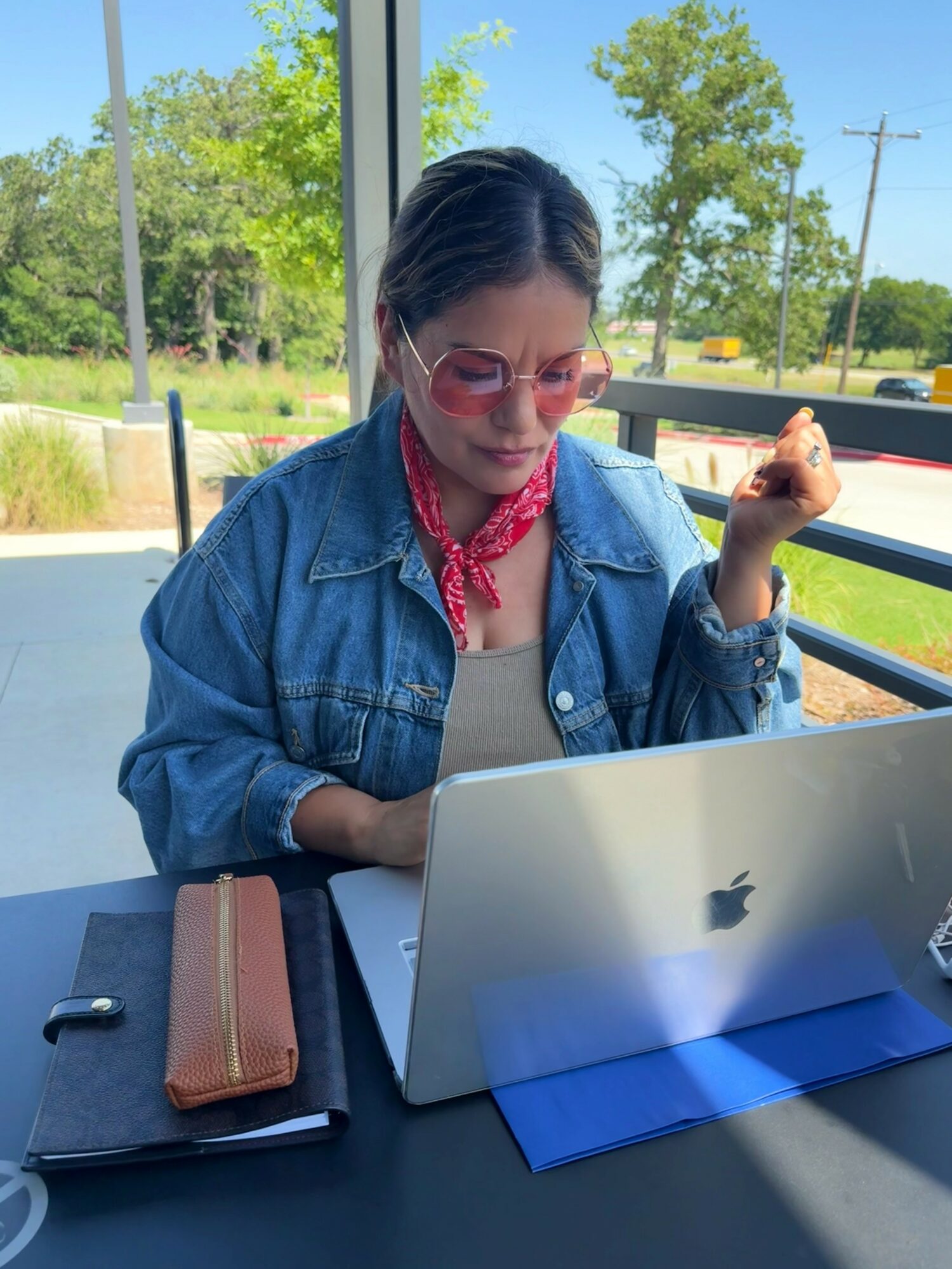 Woman with sunglasses and red bandana working on laptop at a table, outdoors with trees and blue sky in background.