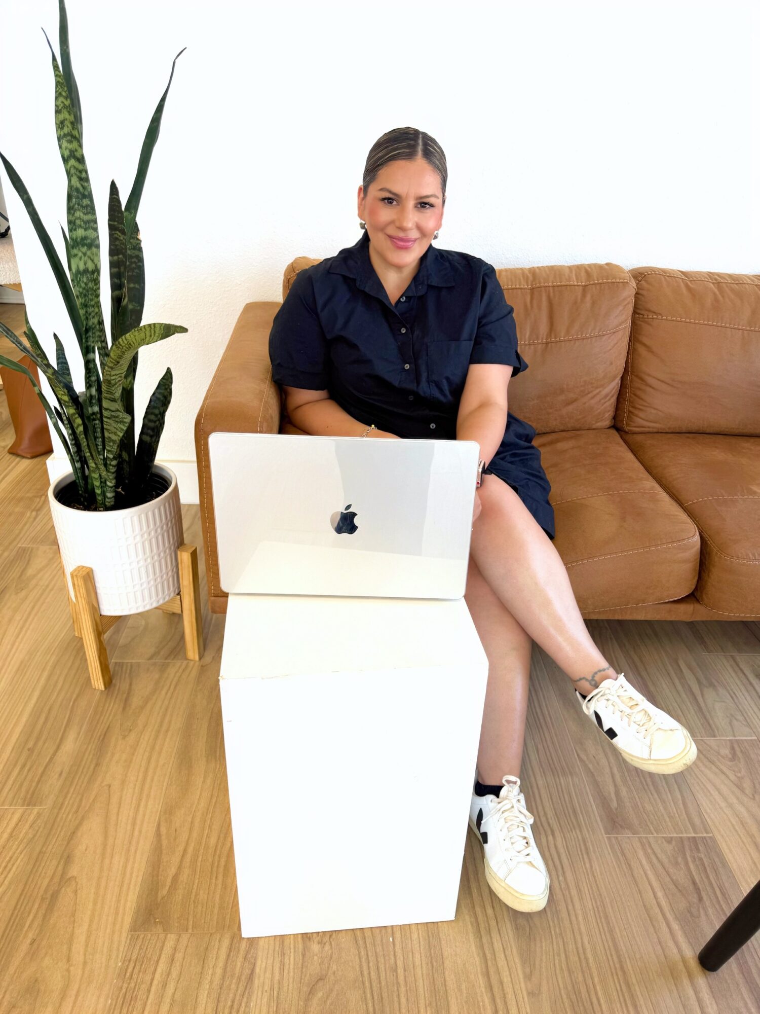 Woman sitting on a sofa with a laptop on a white table, next to a tall potted plant, in a bright room.