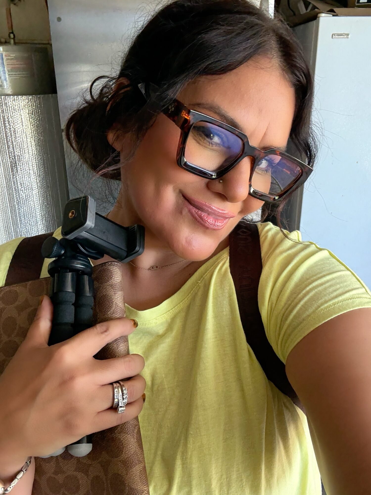Woman with glasses and a yellow shirt smiling, holding a camera, indoors, with a refrigerator in the background.