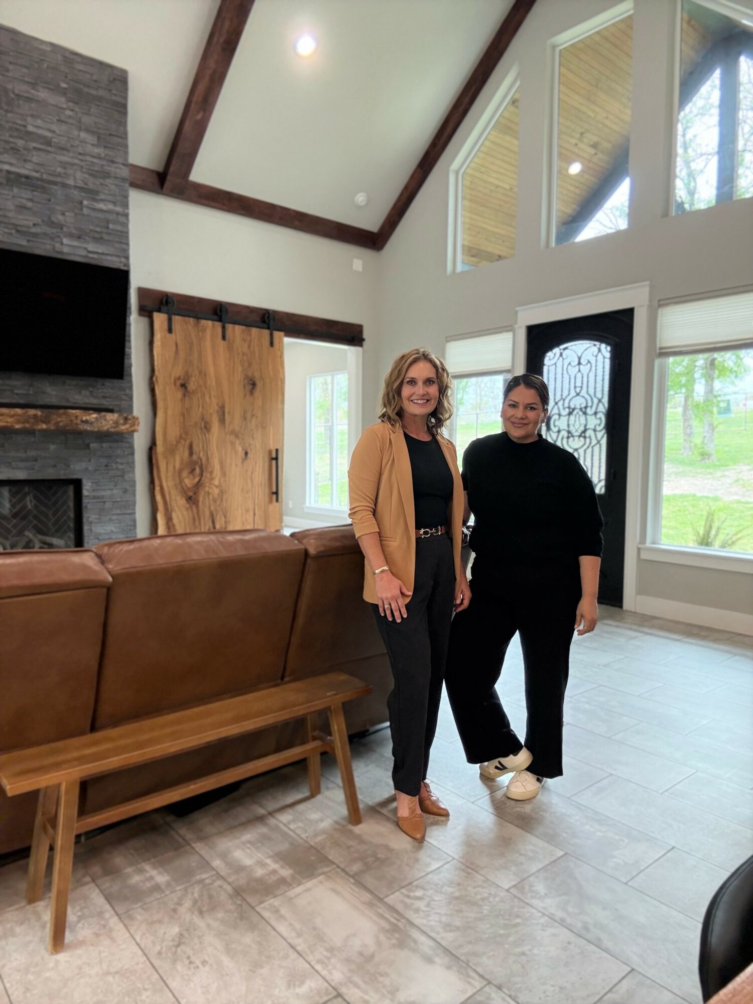 Two women stand inside a spacious, modern living room with large windows, a wooden bench, and a stone fireplace.