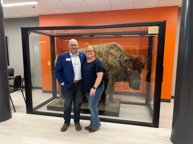 Two people stand inside a glass display case with a large animal pelt, looking at the camera, in a room with orange and white walls.