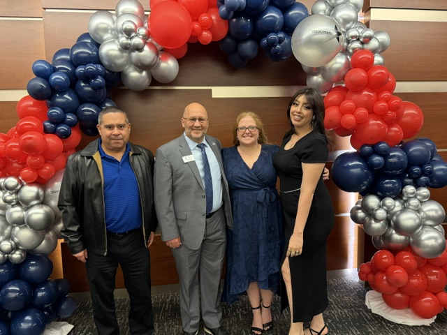 Four people standing in front of a balloon arch with red, blue, and silver balloons, smiling at the camera.