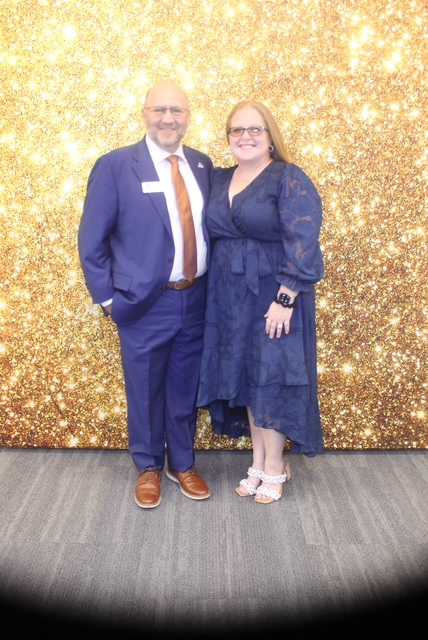 A man and woman stand together in front of a gold glittery backdrop, smiling, dressed in formal attire.