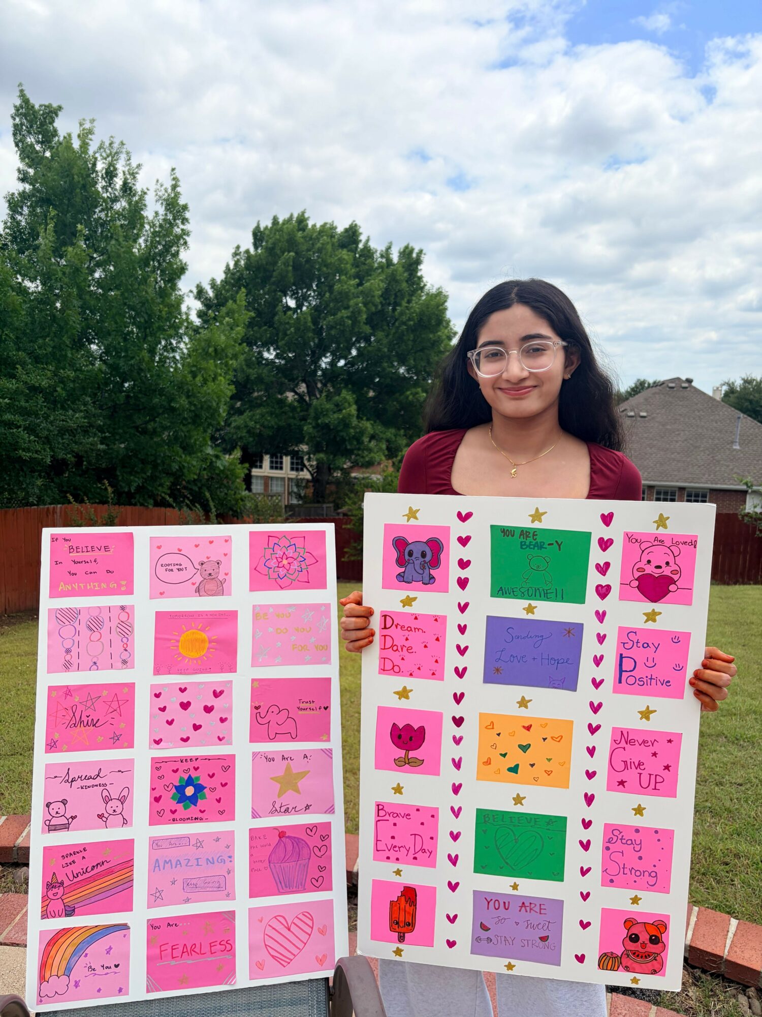 Woman holding colorful handmade cards outdoors with trees and cloudy sky in background.