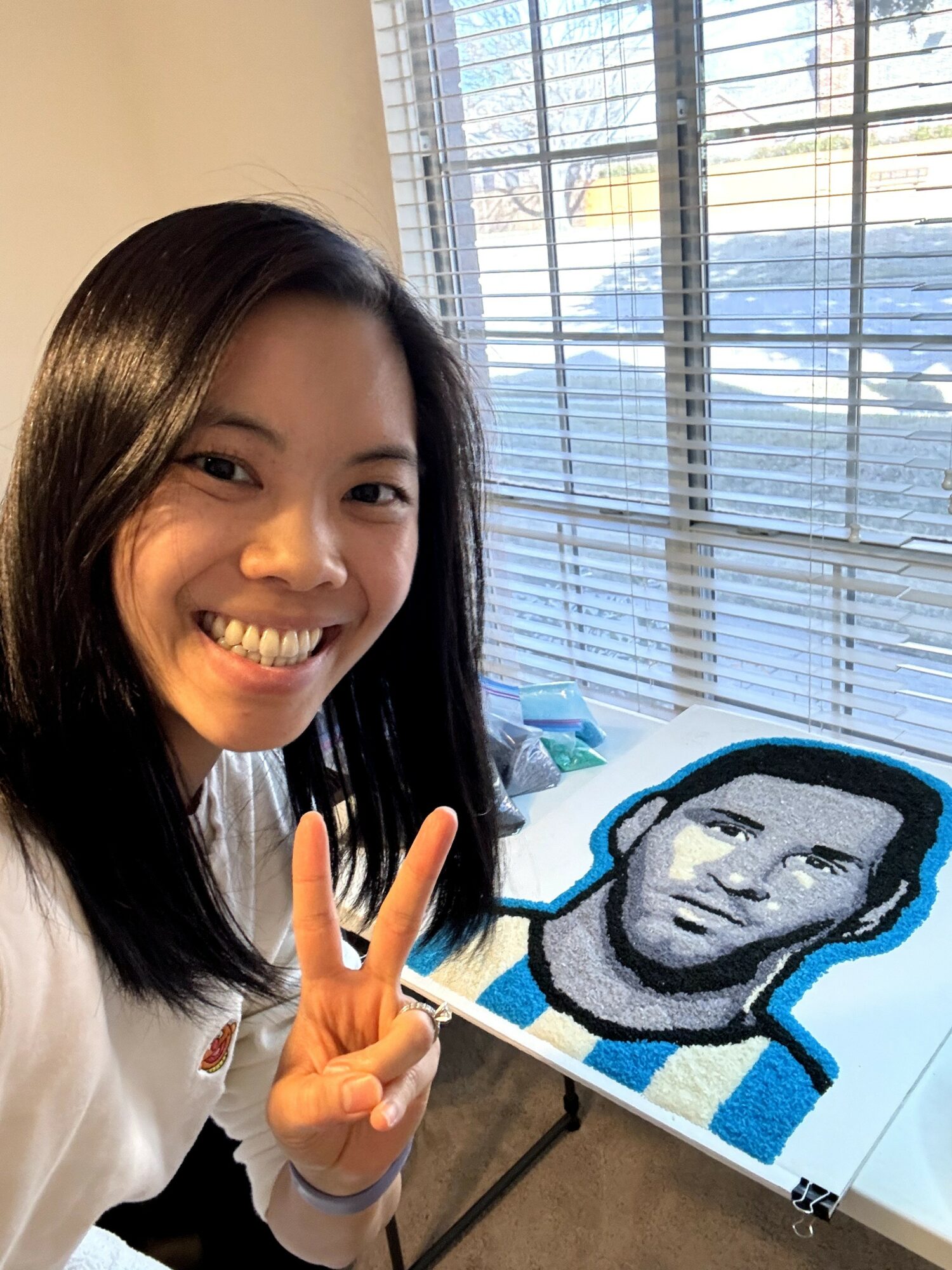 Young woman smiling and making a peace sign next to a pixel art portrait on a table, with window blinds behind.