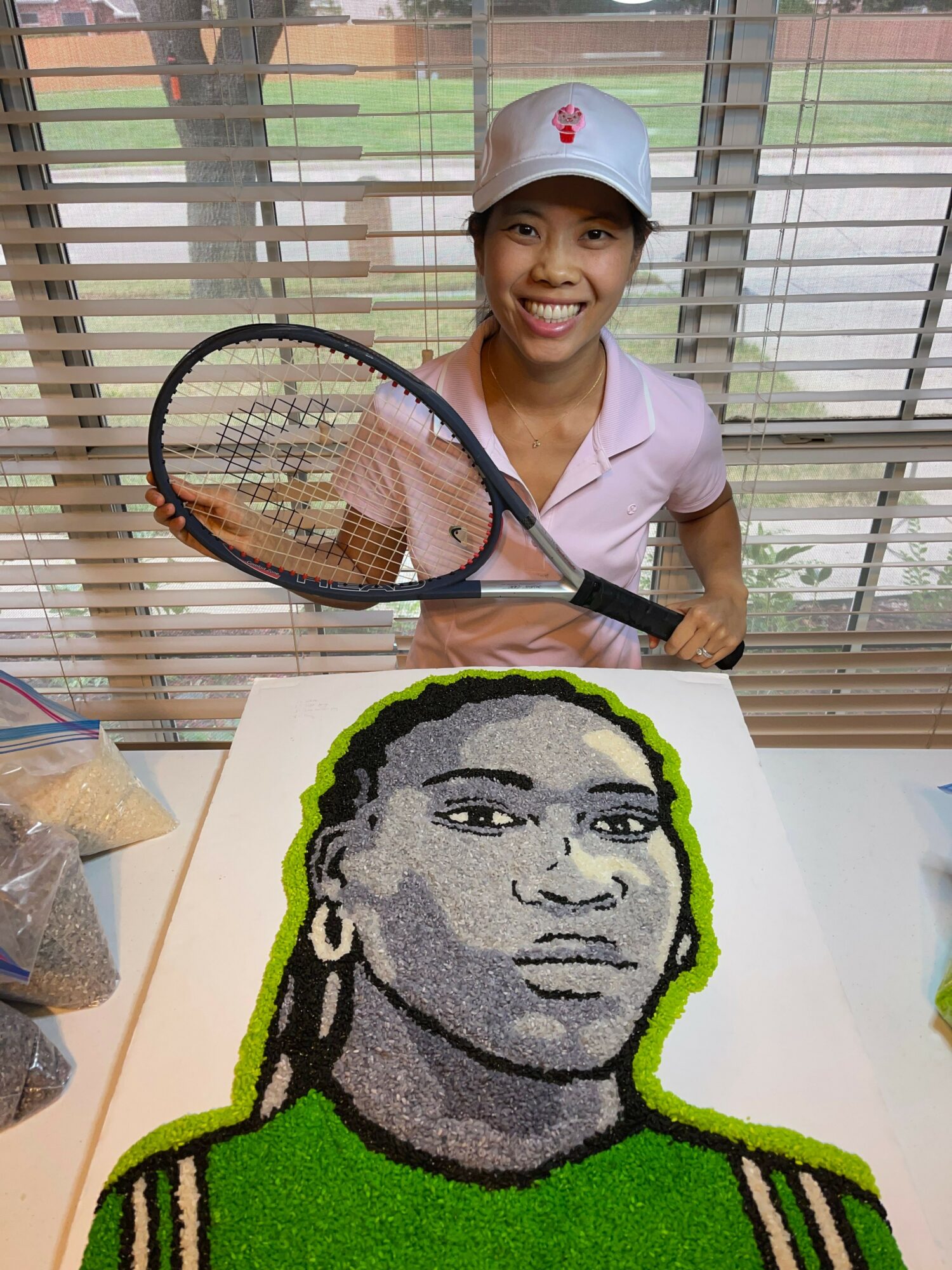 Female tennis player in white cap and pink shirt holding a tennis racket, standing next to a large pixel art portrait of a woman with braided hair, on a white background.