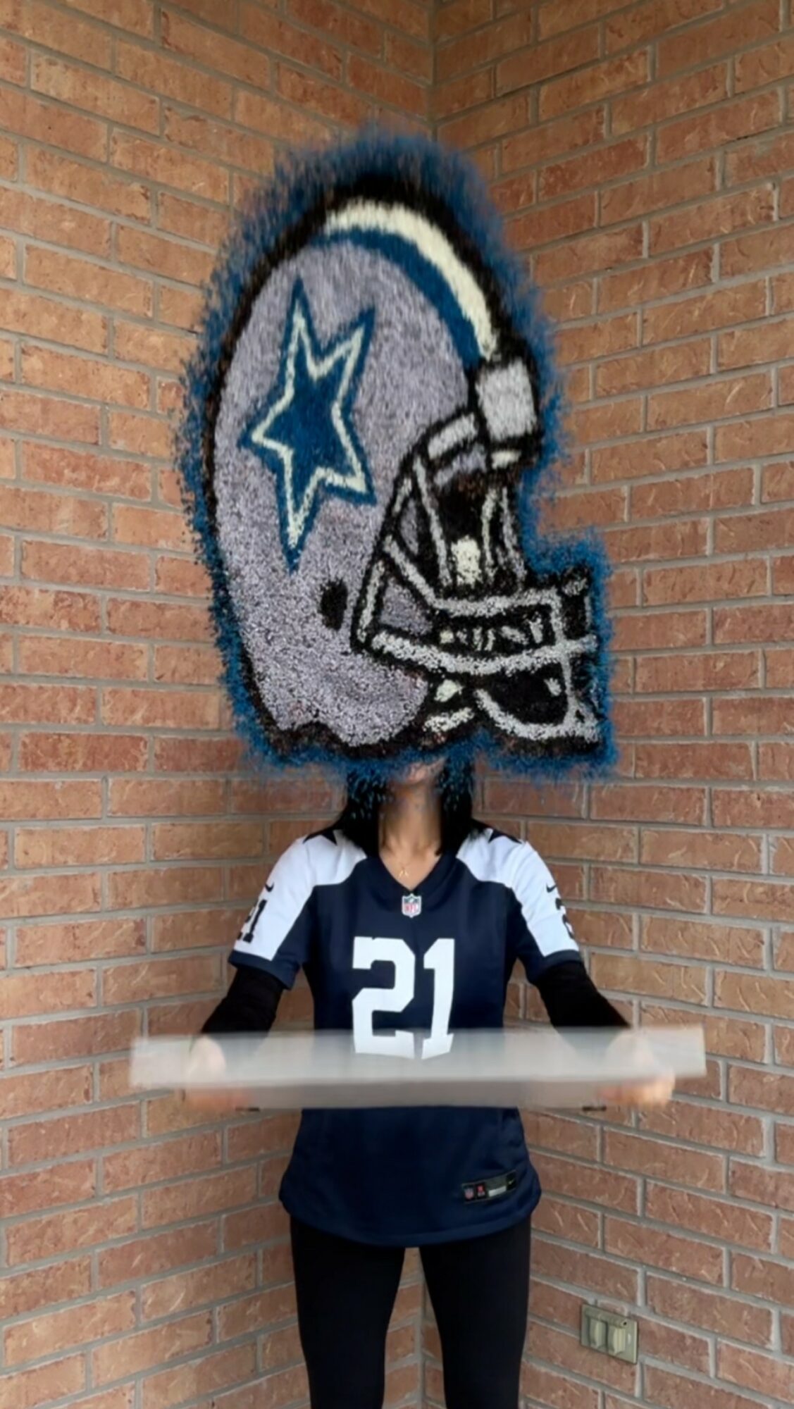 Person wearing a Dallas Cowboys jersey with number 21, holding a table, standing in front of a brick wall with a large Dallas Cowboys helmet mural.