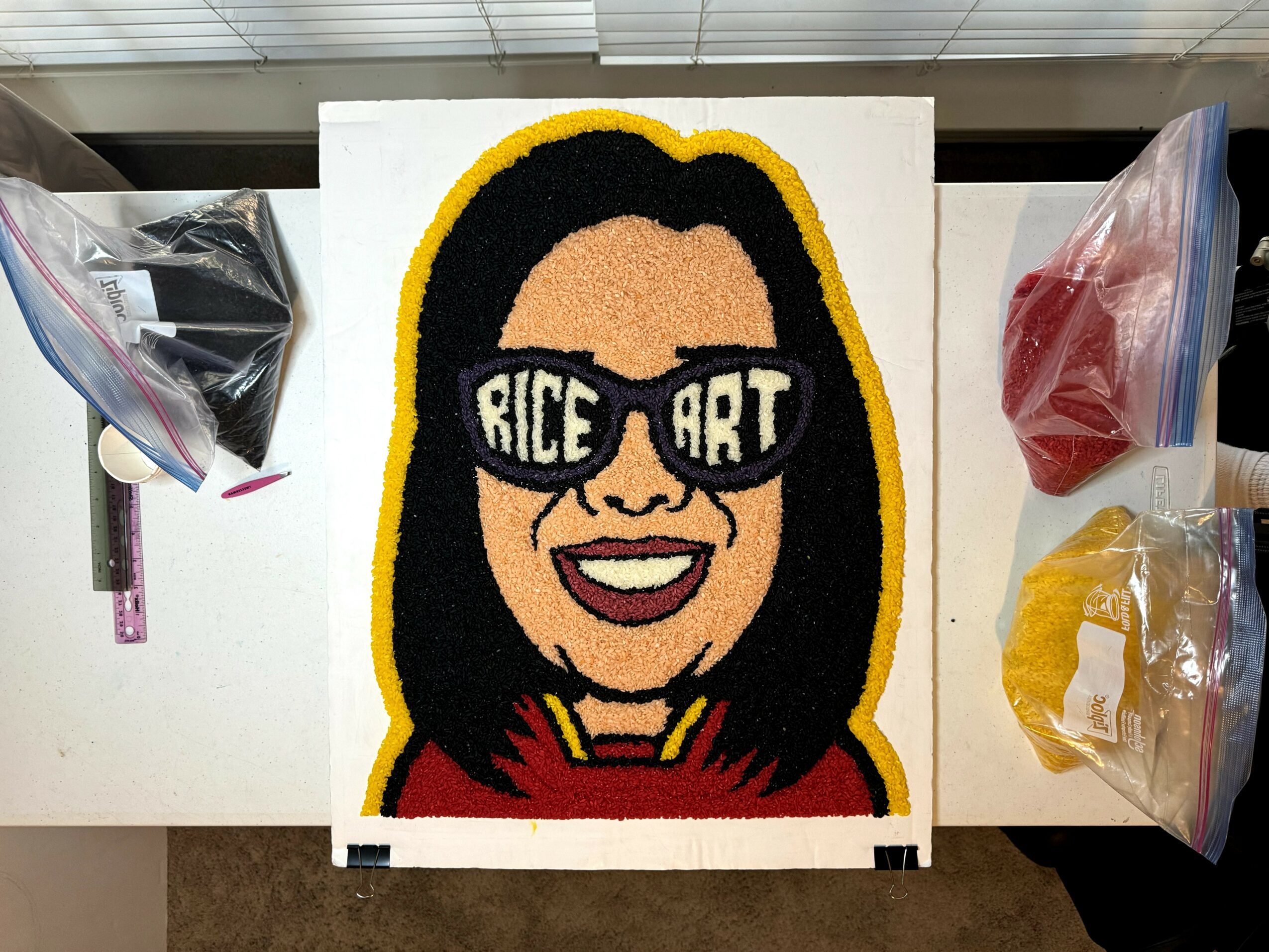 Colorful flower art of a woman with black hair, sunglasses, and a red top, smiling, with a yellow border.