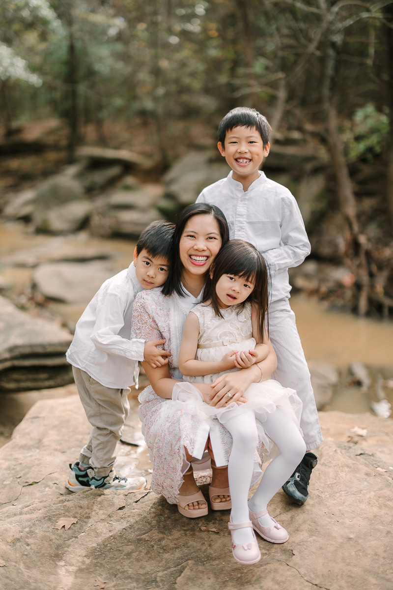 A woman and three children outdoors on rocks, smiling, with trees and a stream in the background.
