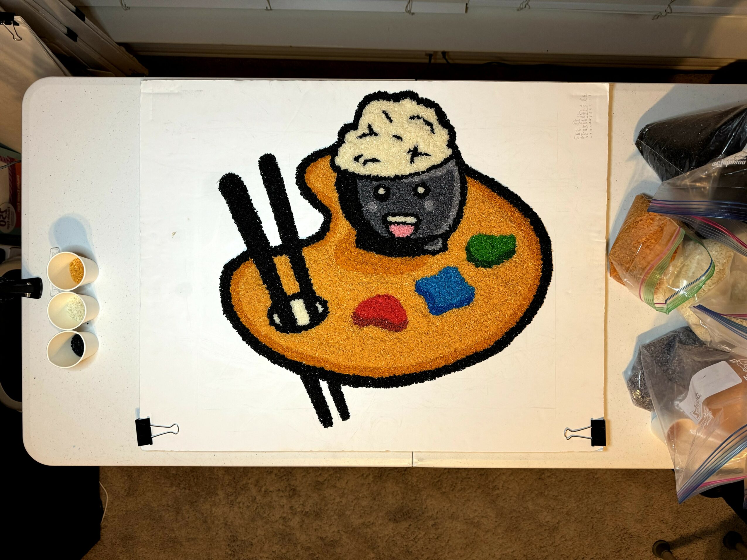 Drawing of a bowl of rice with a smiling face, on a colorful palette with chopsticks, surrounded by art supplies.