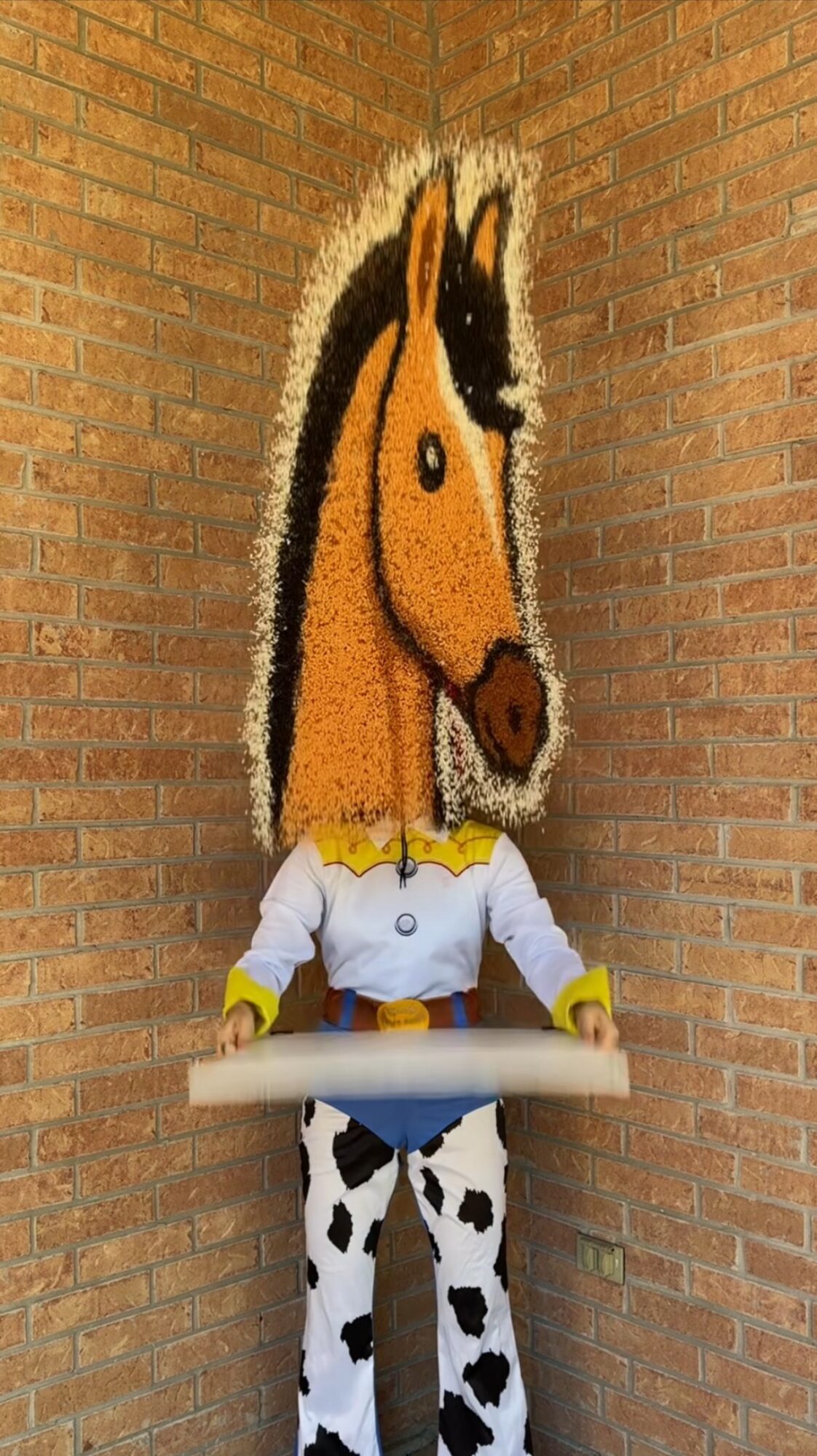 Person in a horse costume with a large horse head, standing against a brick wall, holding a white object.