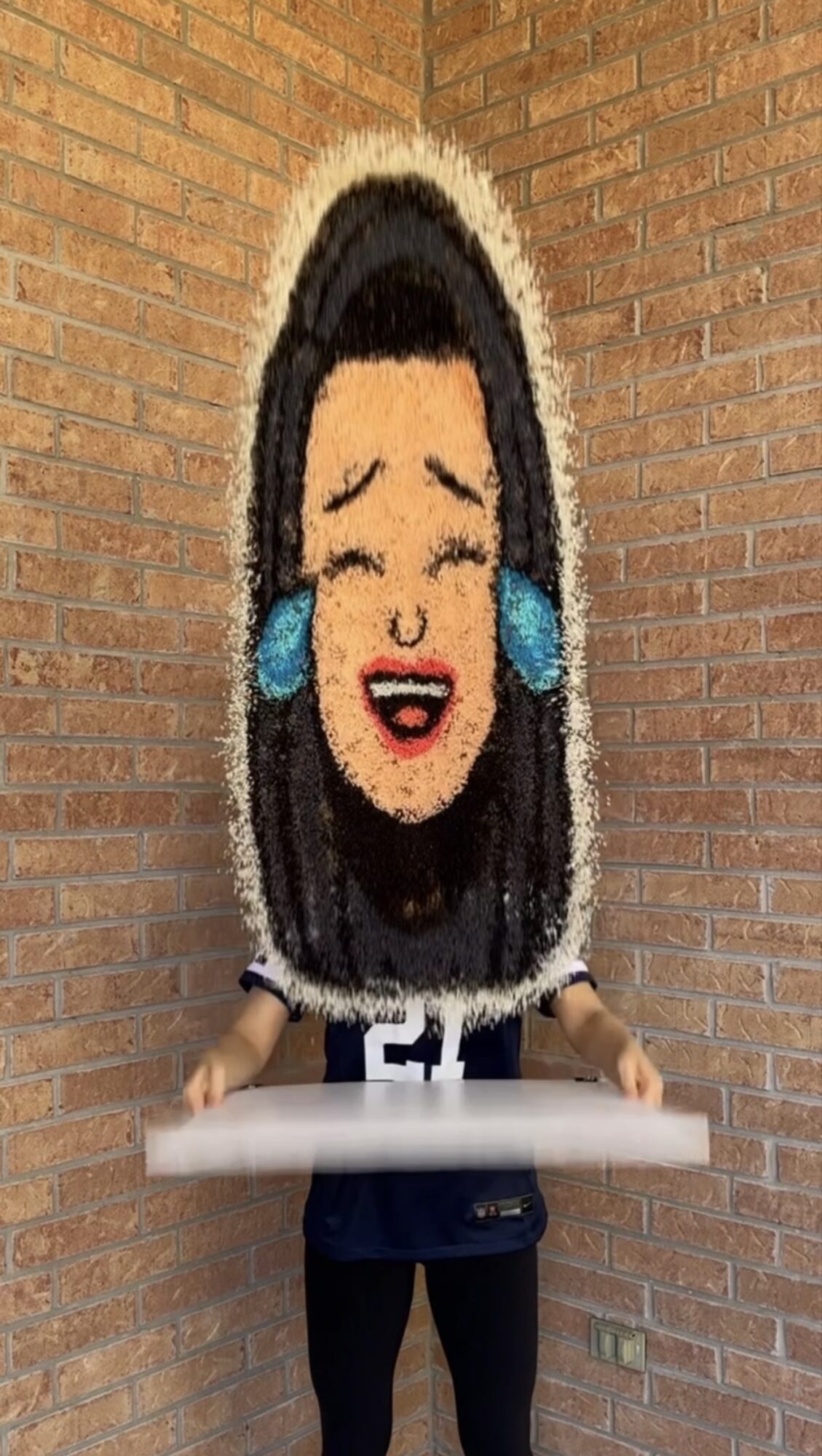 Person holding a large, colorful face mask with a smiling face and blue earrings, standing against a brick wall.