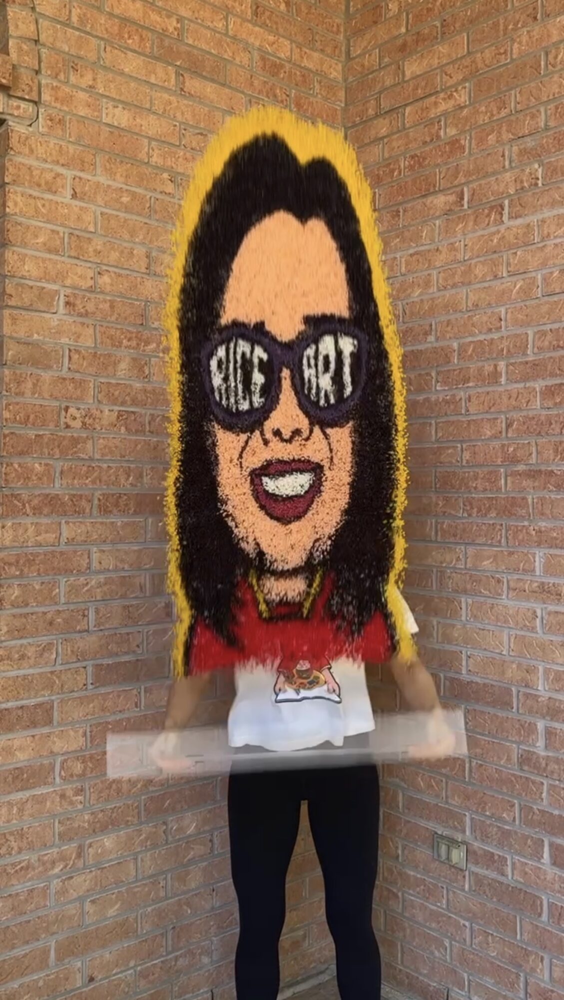 Person holding a large pixel art portrait of a woman with sunglasses and dark hair, standing against a brick wall.