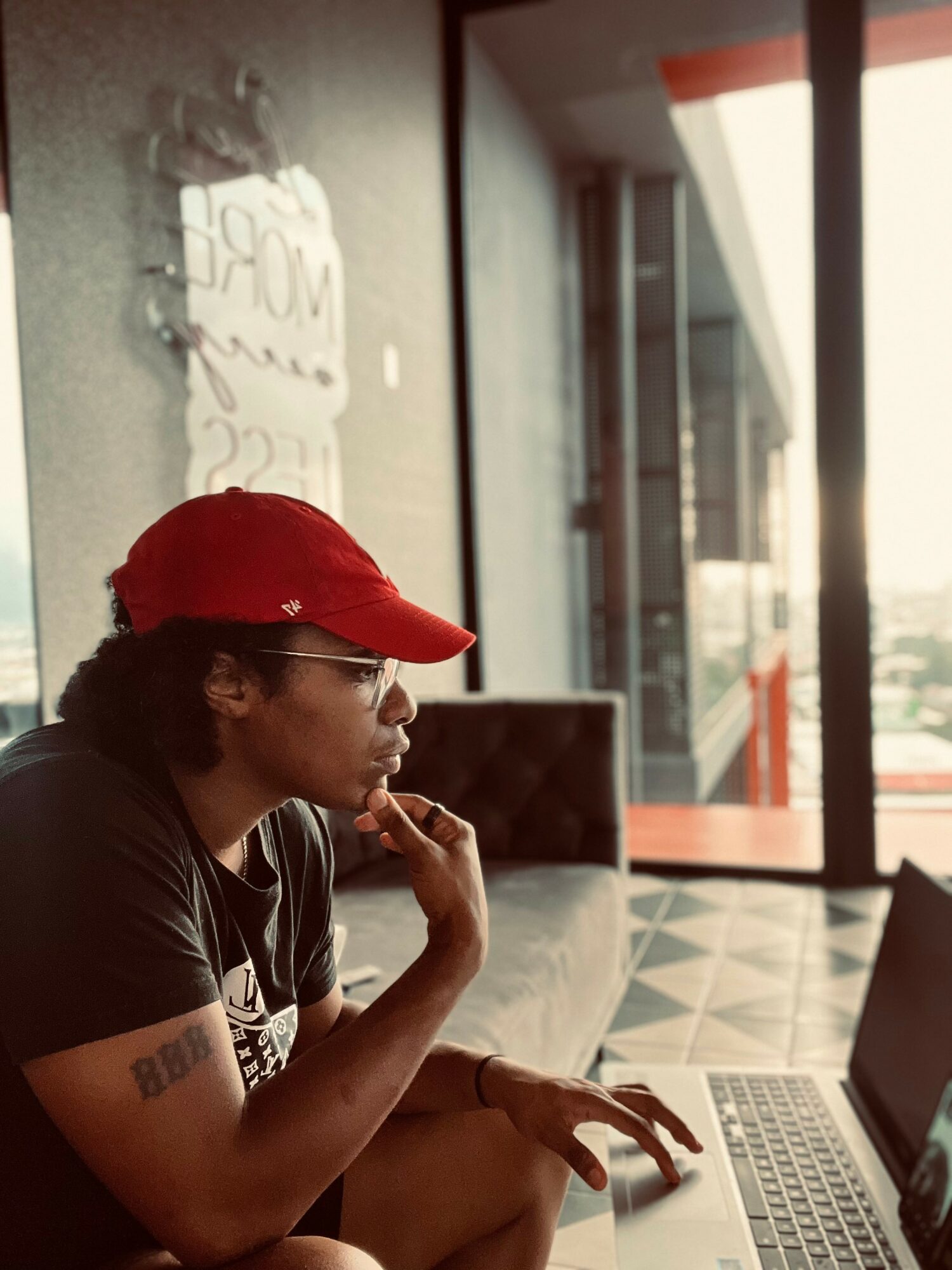 Person wearing a red cap and glasses, sitting at a table with a laptop, in a modern indoor space with large windows.