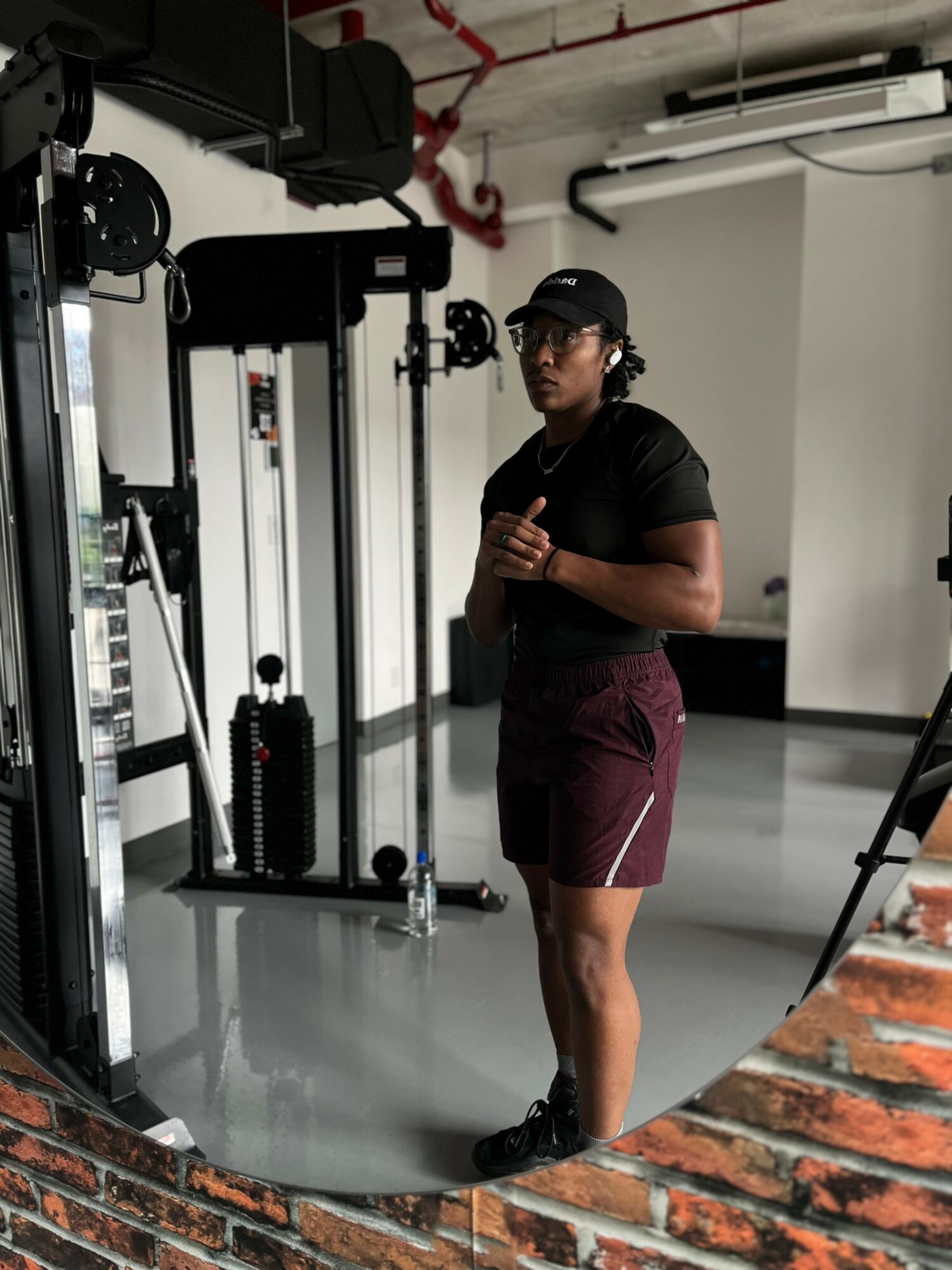Person in gym wearing black shirt, maroon shorts, and cap, standing near exercise equipment, looking at the camera.