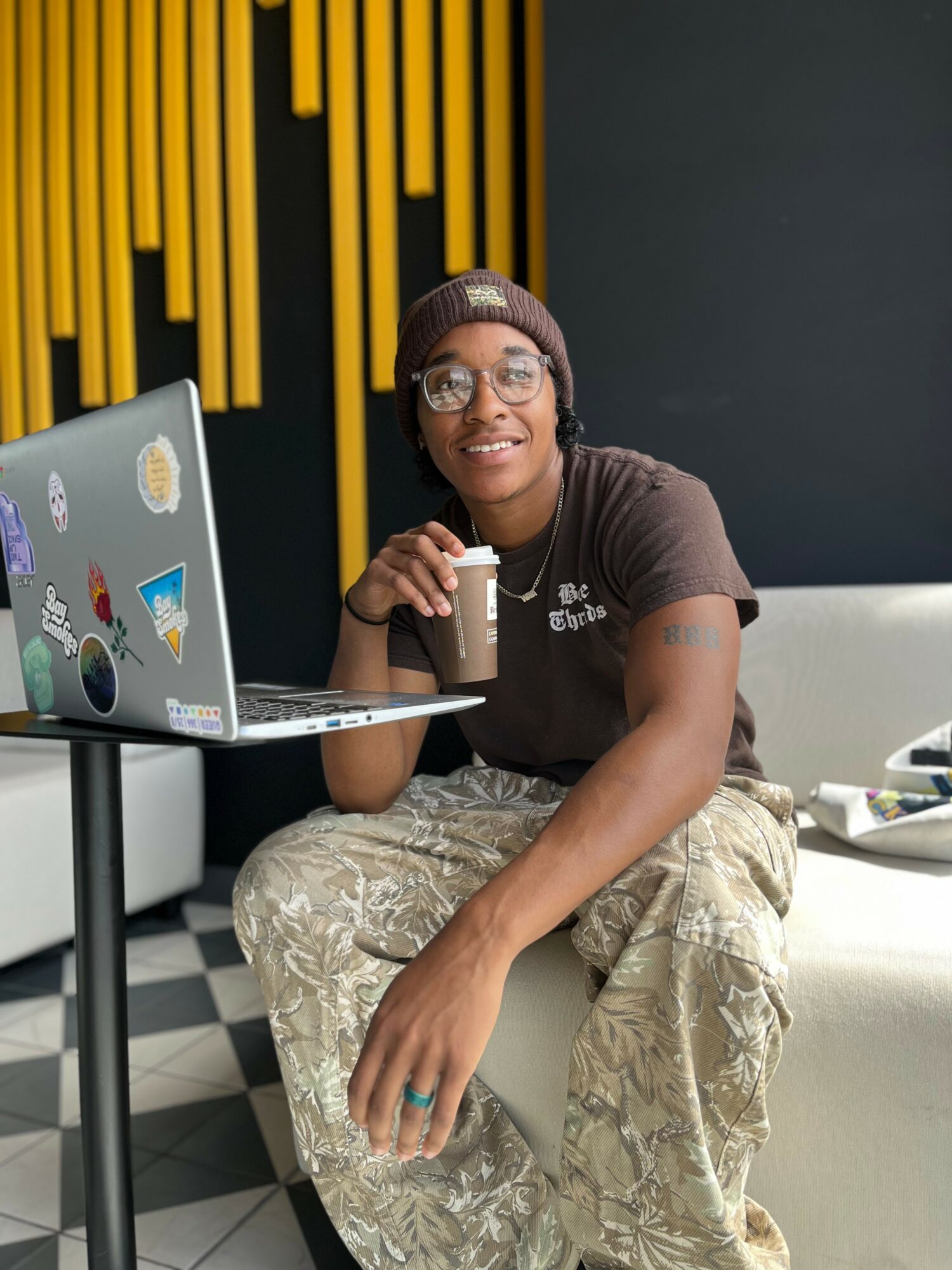 Person sitting on a white couch with a laptop decorated with stickers, holding a drink, smiling, in a modern room.