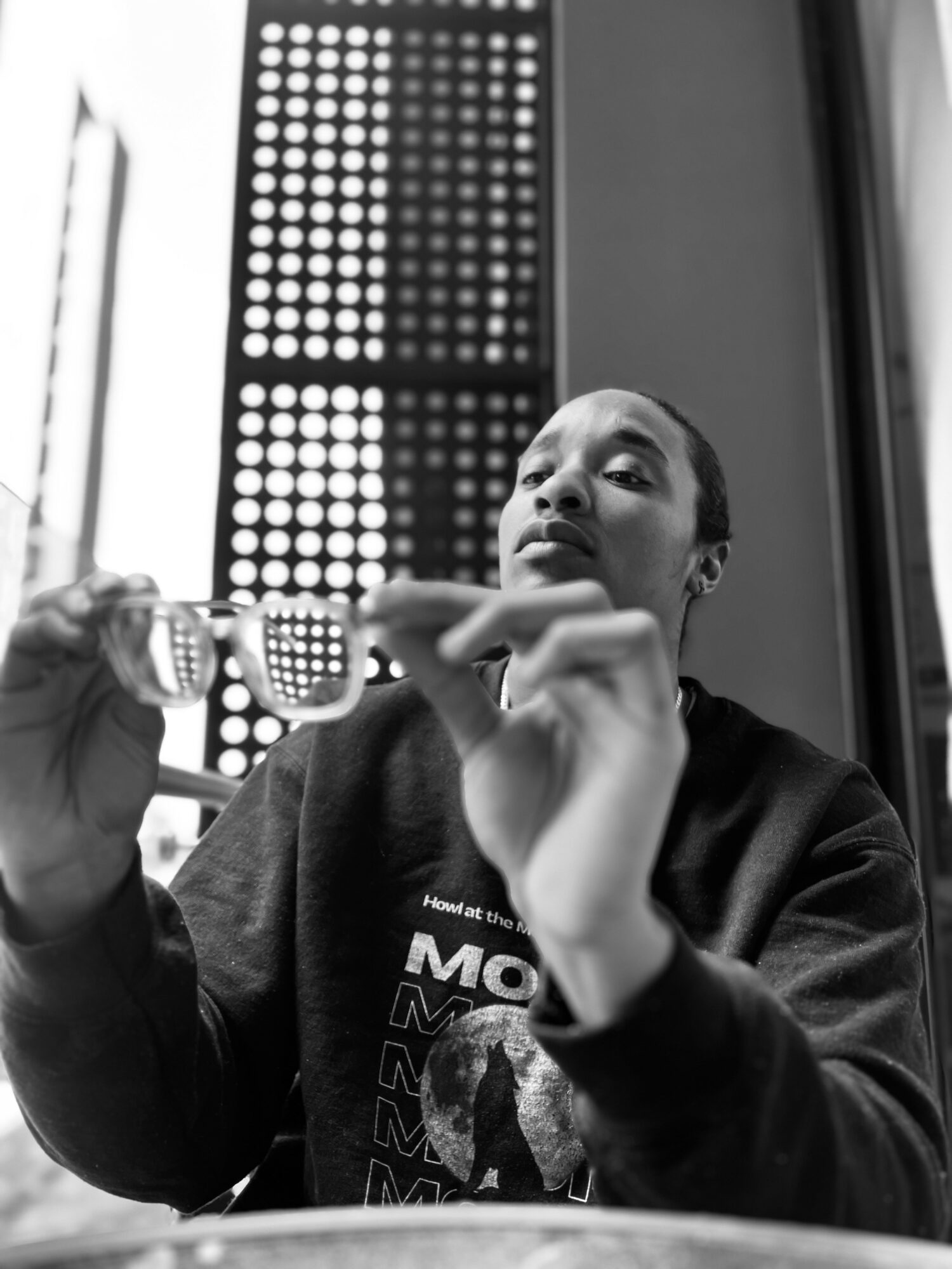 Person holding glasses, looking at camera, indoors with patterned window behind, in black and white.