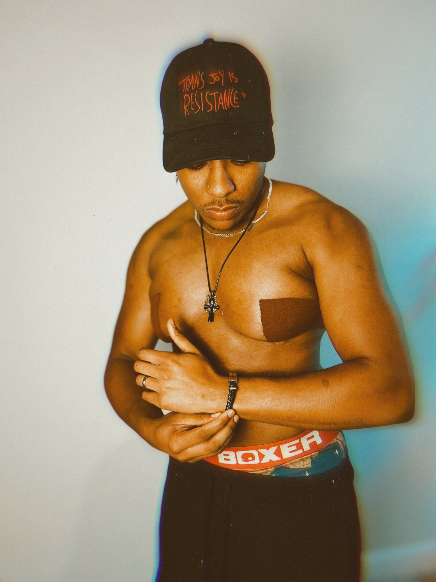 Young shirtless man wearing a black cap with red text, black pants, and a cross necklace, standing against a plain background.