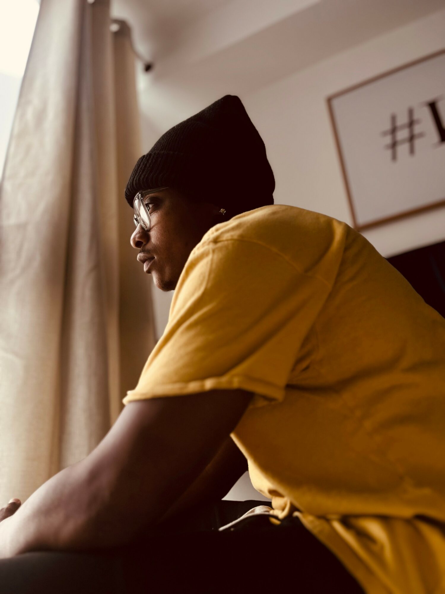 Person wearing glasses, black beanie, and yellow shirt sitting indoors near a window with curtains.
