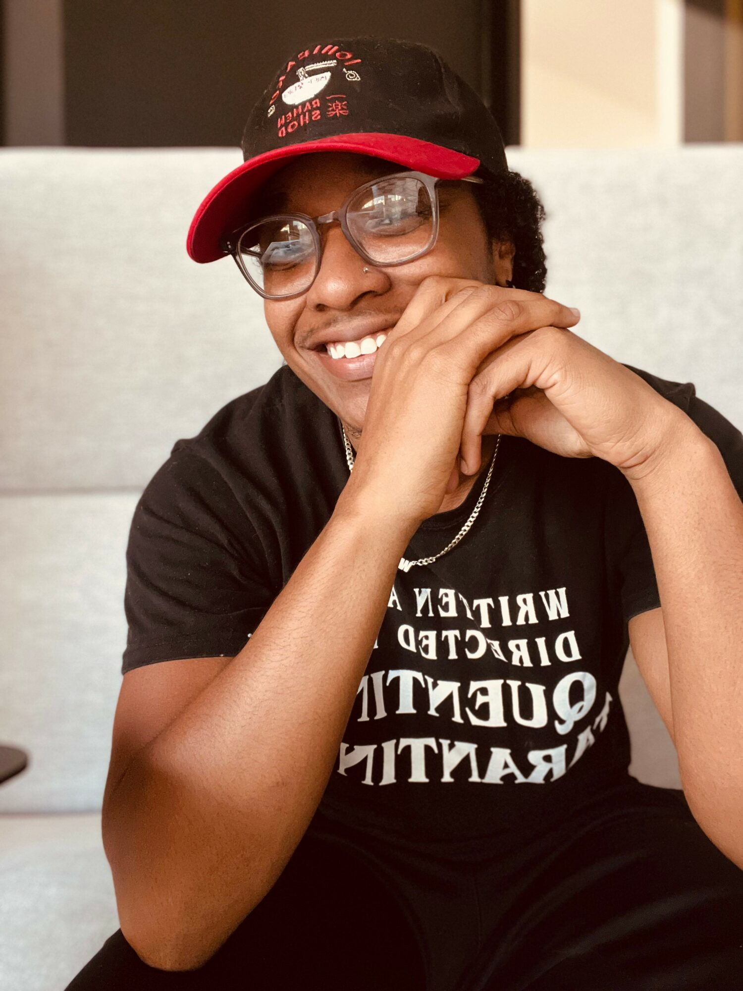 Person smiling, wearing glasses and a black and red cap, resting chin on hand, indoors.