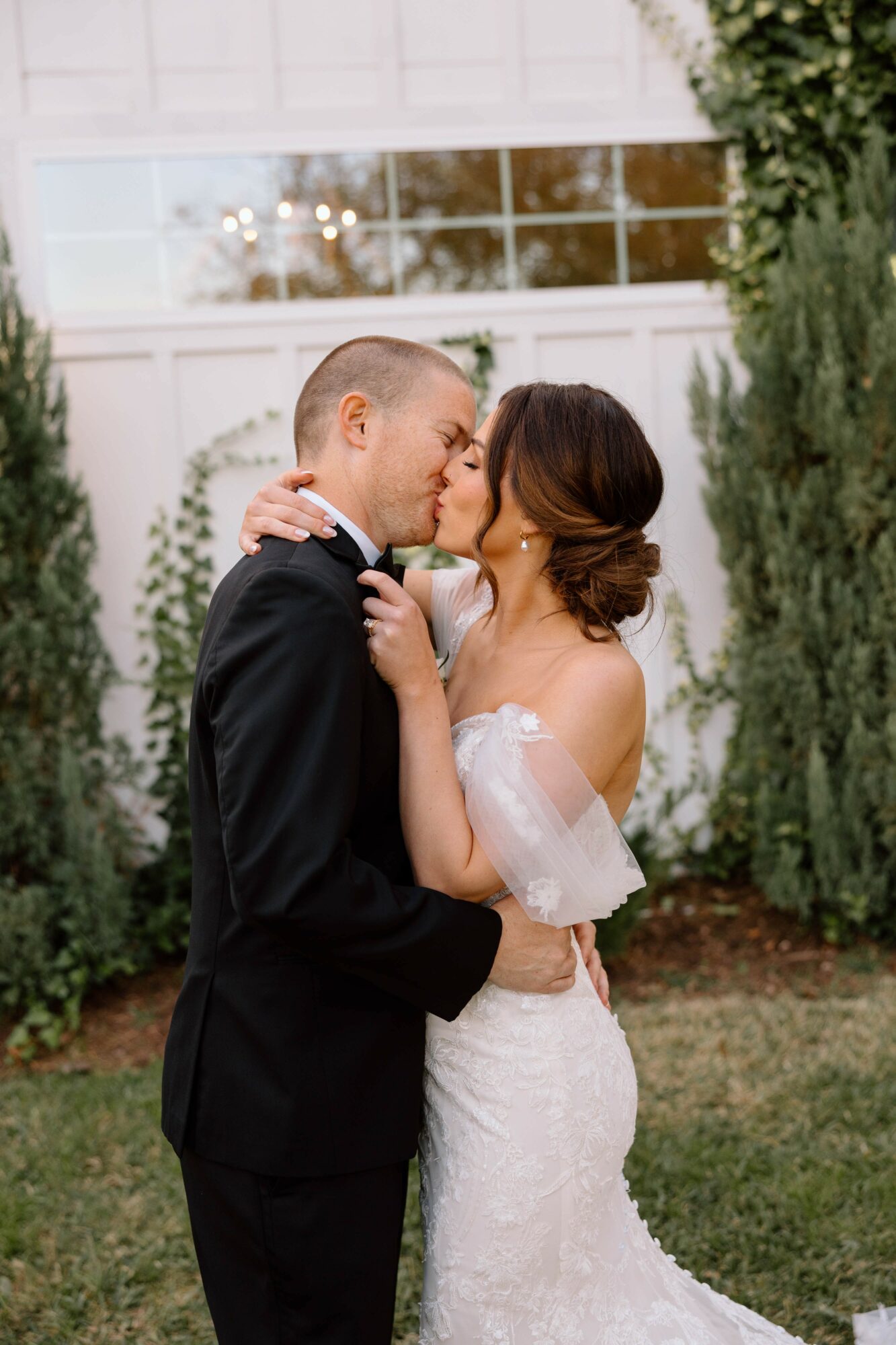 Bride and groom kiss outdoors, surrounded by greenery and a white fence, during daytime.