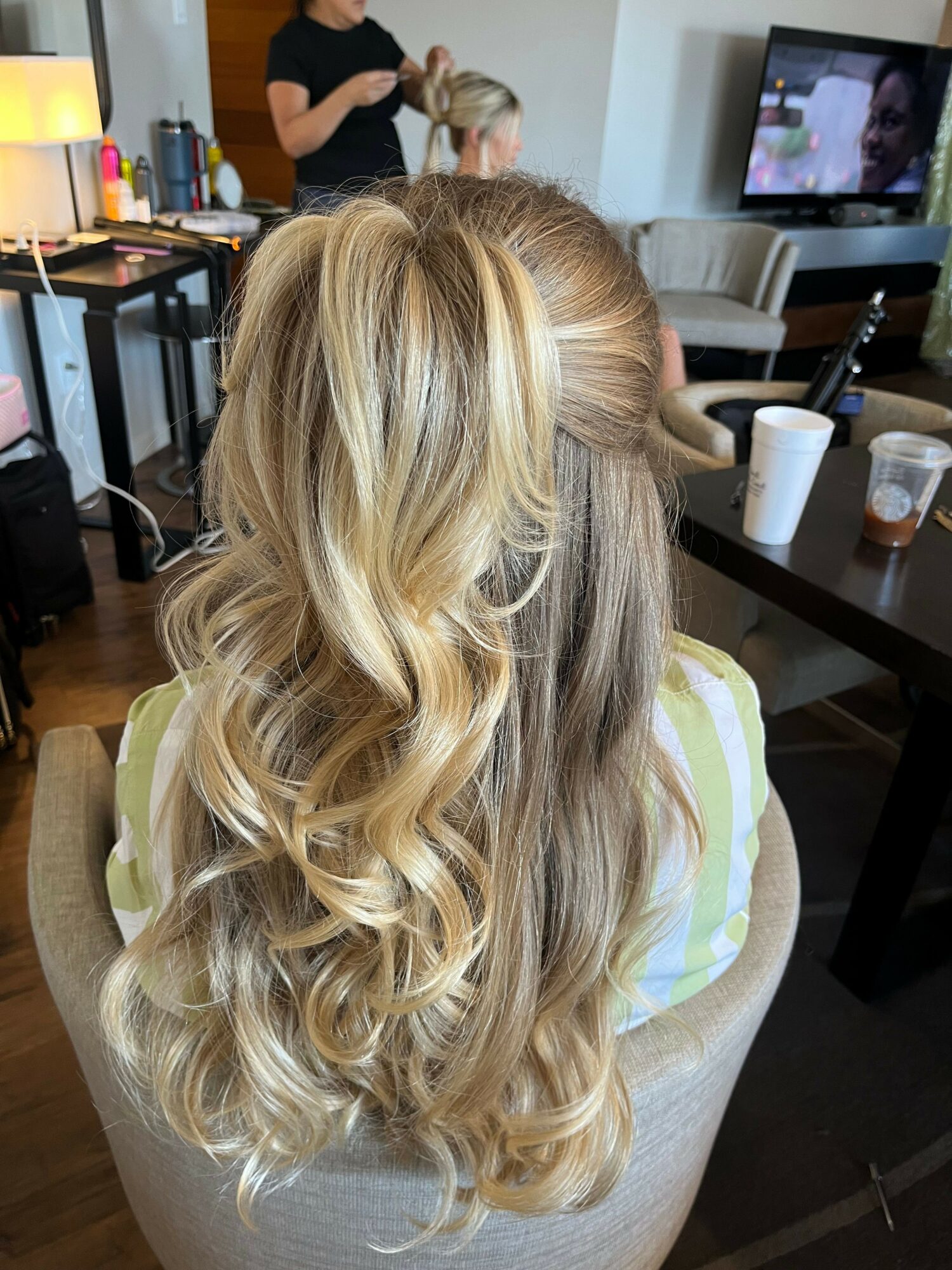 Person with long, wavy blonde hair sitting in a salon chair, viewed from behind, with salon tools and a mirror in the background.