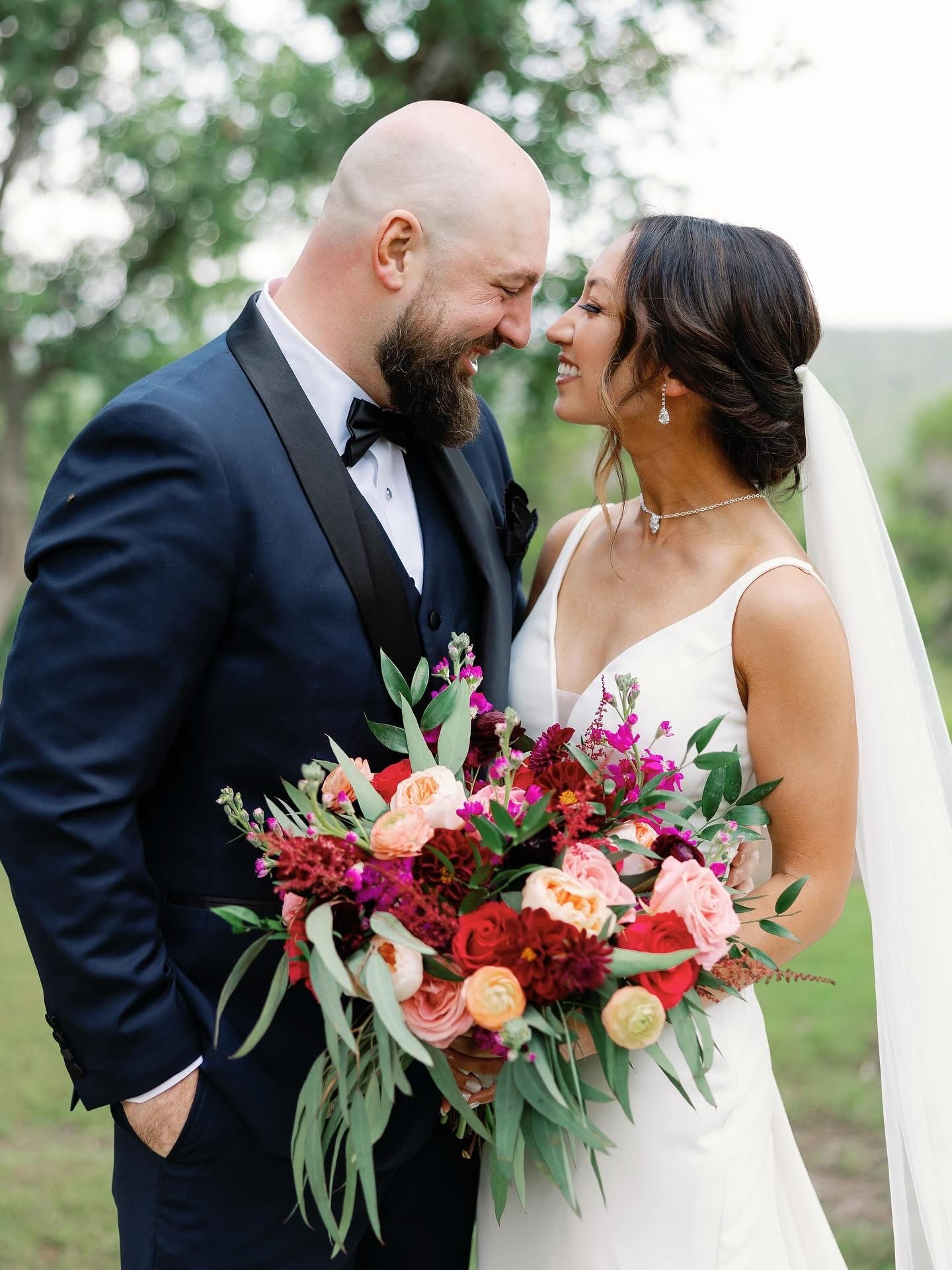 Bride and groom face each other outdoors, smiling, with the bride holding a large colorful bouquet, trees in background.