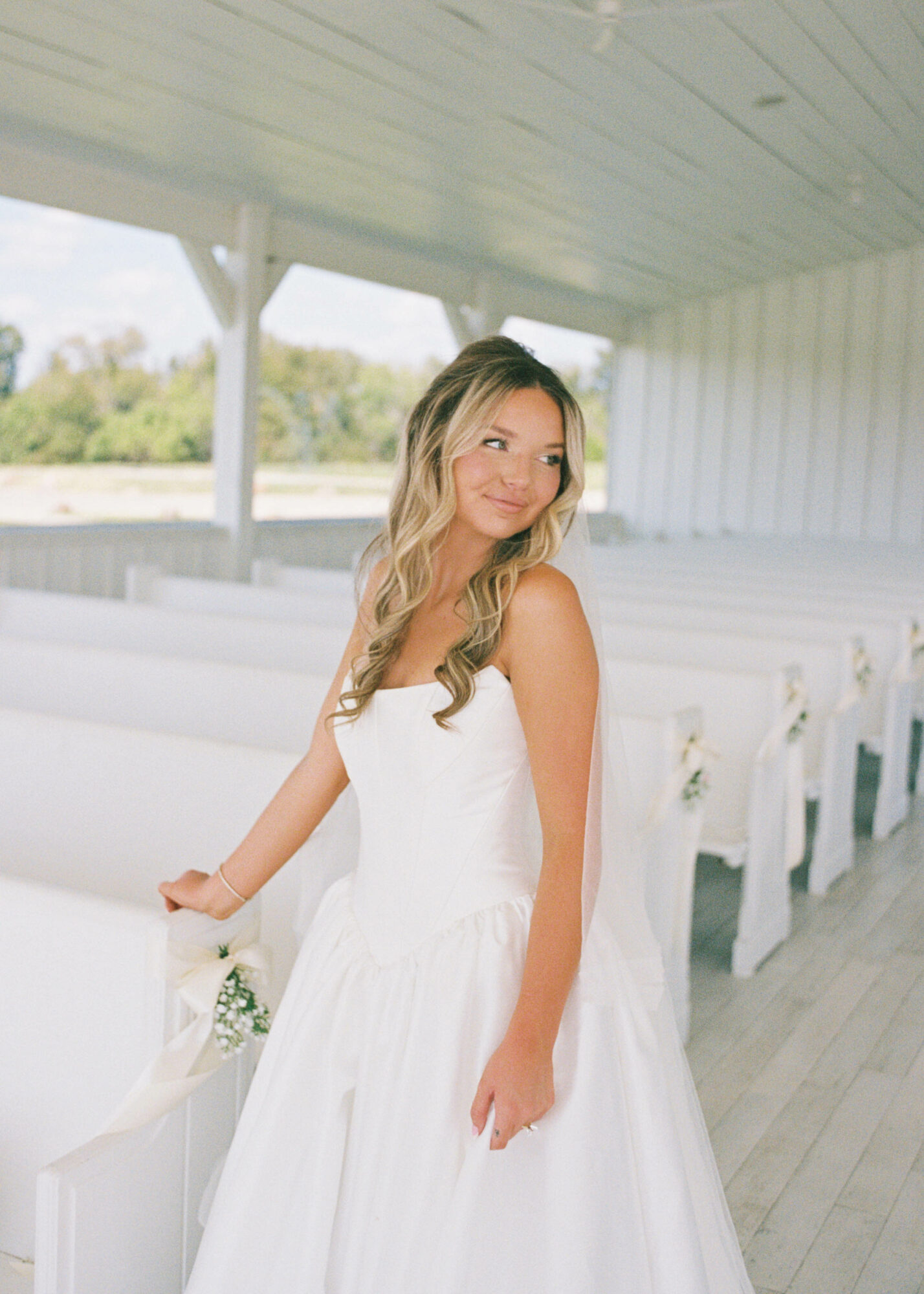 Young woman in a white dress standing inside a bright, open space with white benches and a view outside.
