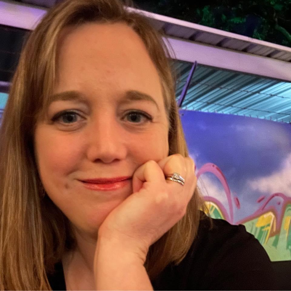 Close-up of a woman with light brown hair resting her chin on her hand, smiling softly, with a colorful background.