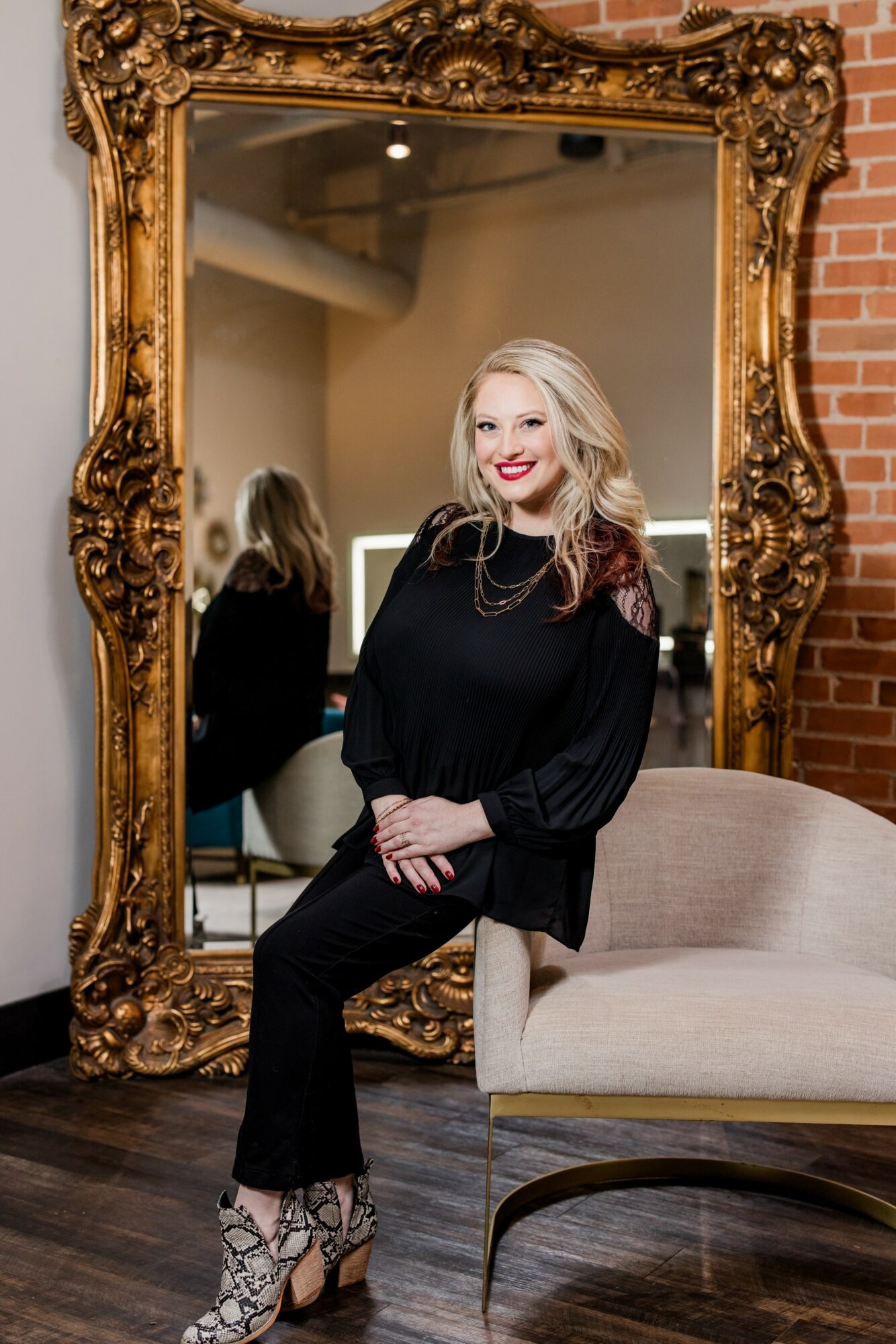 Woman with blonde hair and red lipstick sitting on a beige sofa, reflected in an ornate gold mirror, brick wall background.