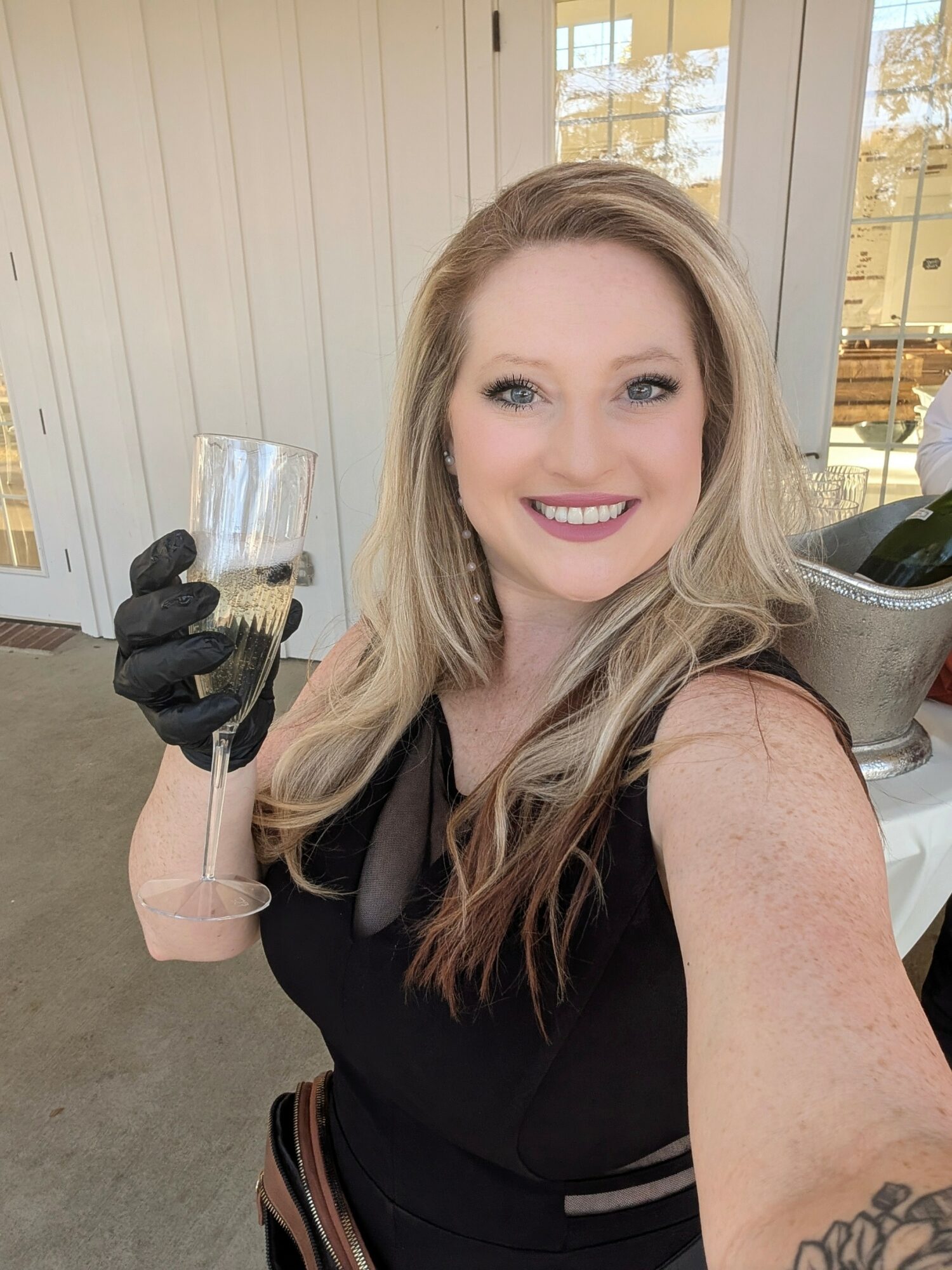 Woman with long blonde hair smiling, holding a glass, wearing a black dress and black gloves, indoors with windows behind.