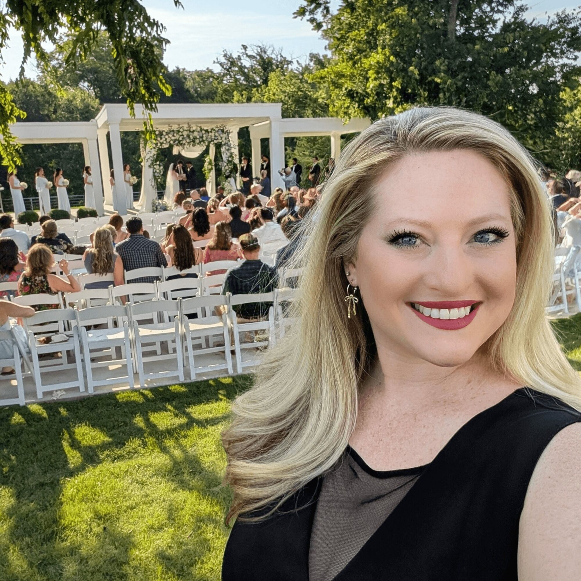 Woman with blonde hair smiling at outdoor wedding ceremony, guests seated, white arch, trees, sunny day.