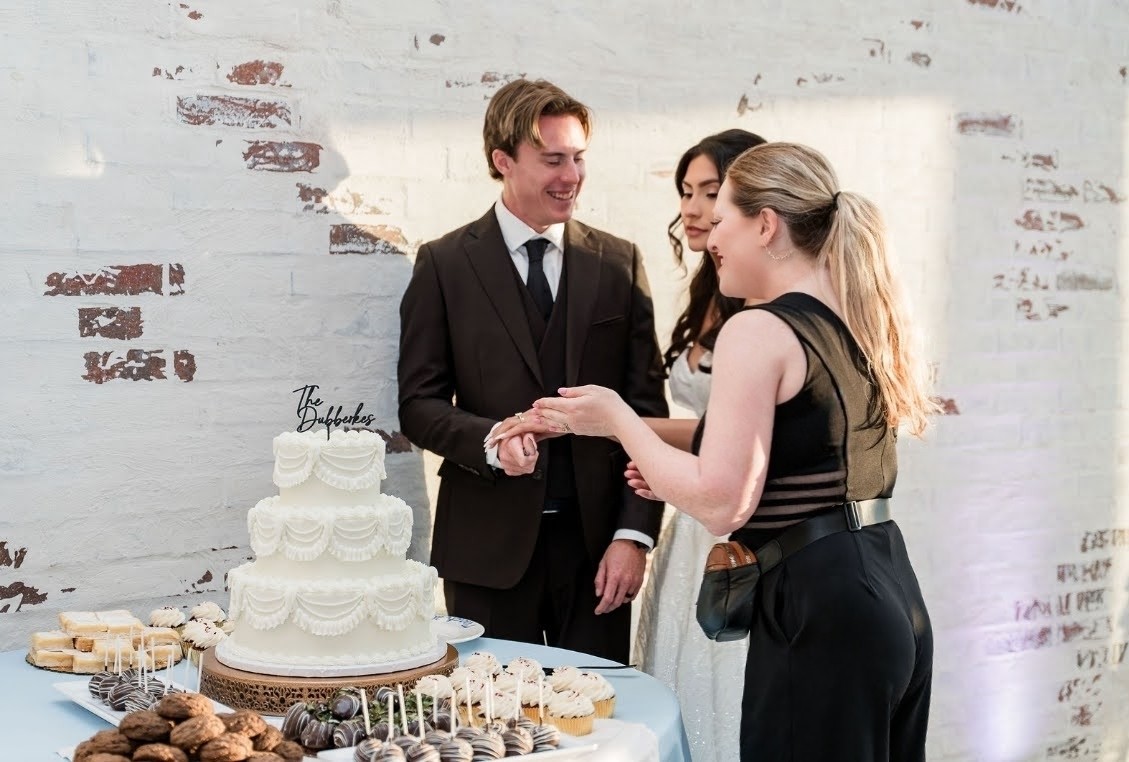 Three people celebrating around a table with a large white cake, holding hands, in front of a white brick wall.