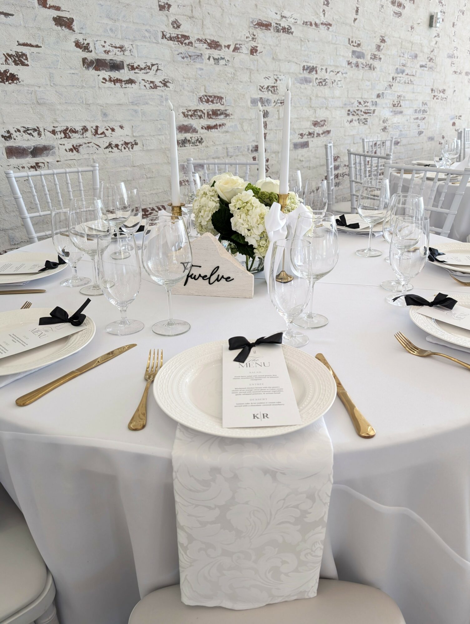 Round table set with white tablecloth, floral centerpiece, glassware, gold cutlery, and black ribbon napkins, against a white brick wall.