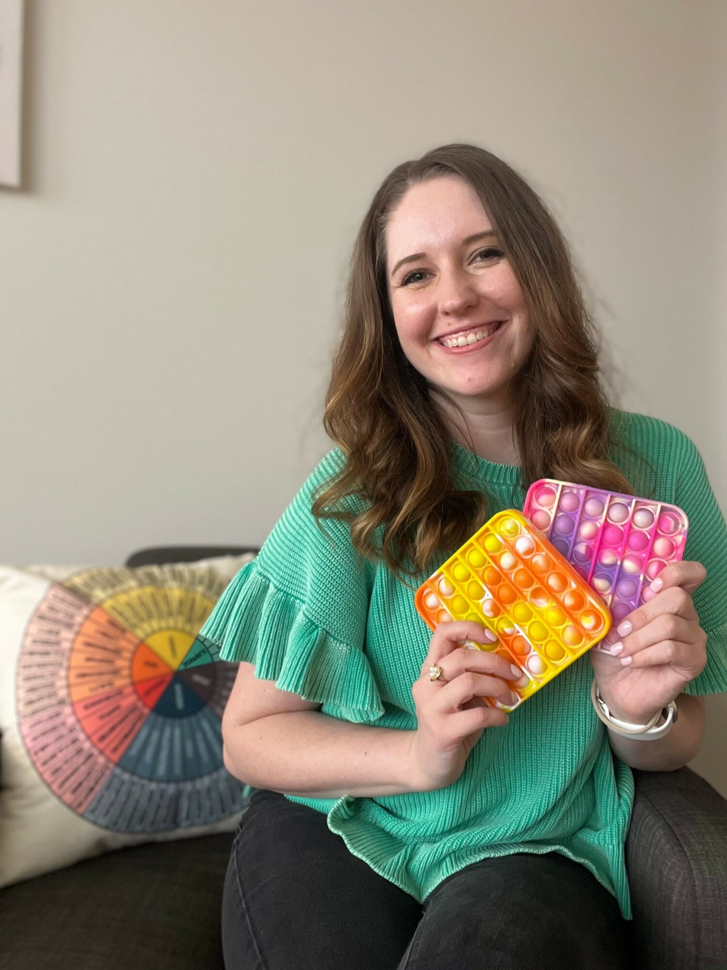 Smiling woman with wavy hair holding colorful pop-it toys, sitting on a couch with a patterned pillow behind her.