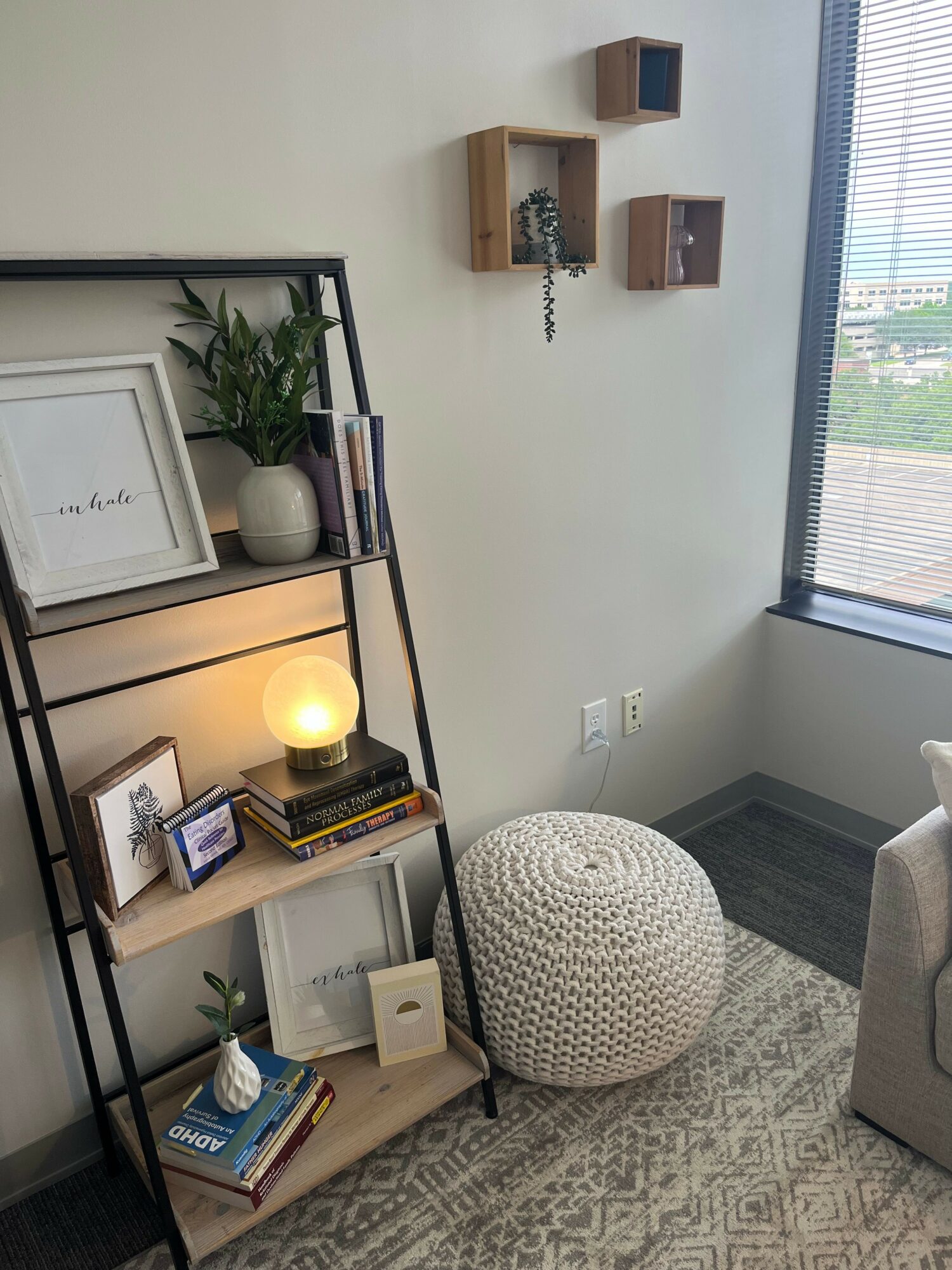 Office corner with a window, wall shelves, a round pouf, and a black metal desk with books and a lamp.